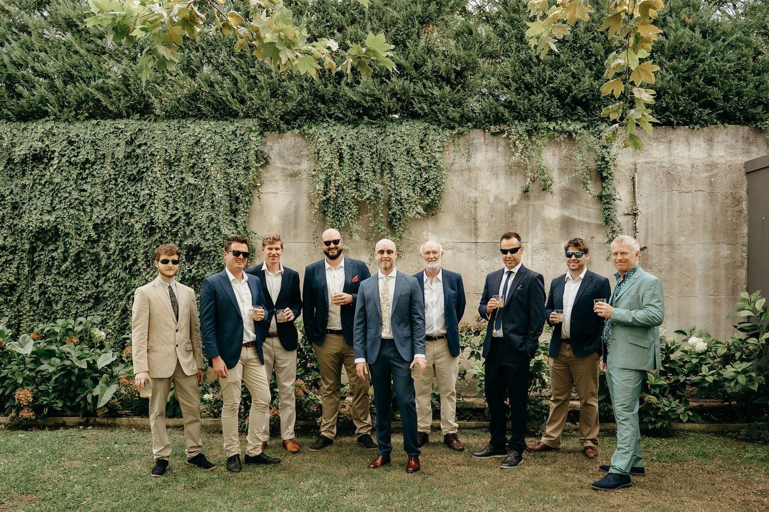 A group of well-dressed men in sunglasses and suits stand together against an ivy-covered wall in a garden setting.