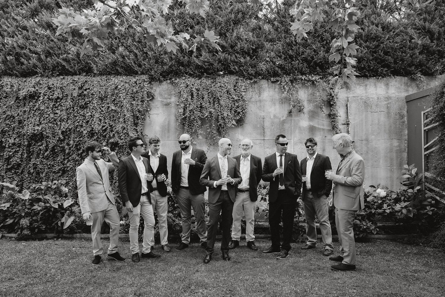 Black and white photo of groomsmen in formal attire standing together in front of a garden wall during a wedding.