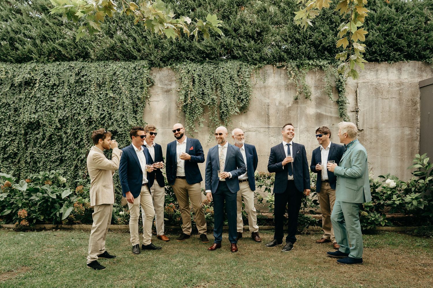 Well-dressed men in formal attire gather for a wedding photo against an ivy-covered concrete wall in a garden setting.