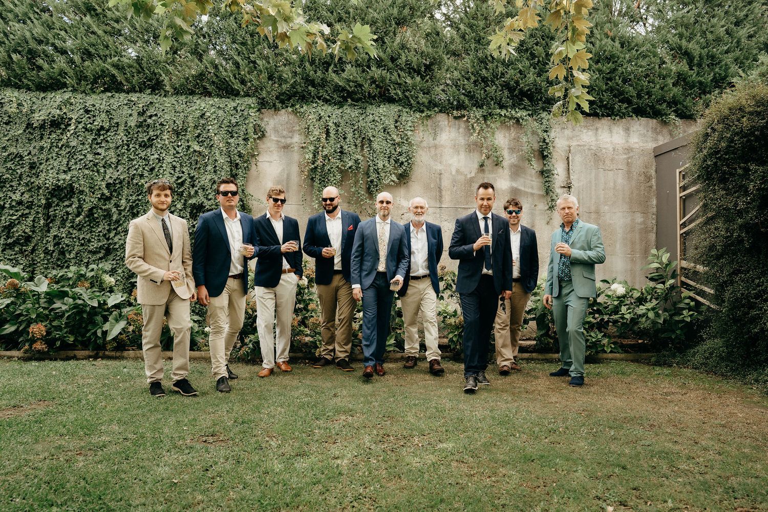 A group of men in formal attire stand together on grass in front of an ivy-covered stone wall.