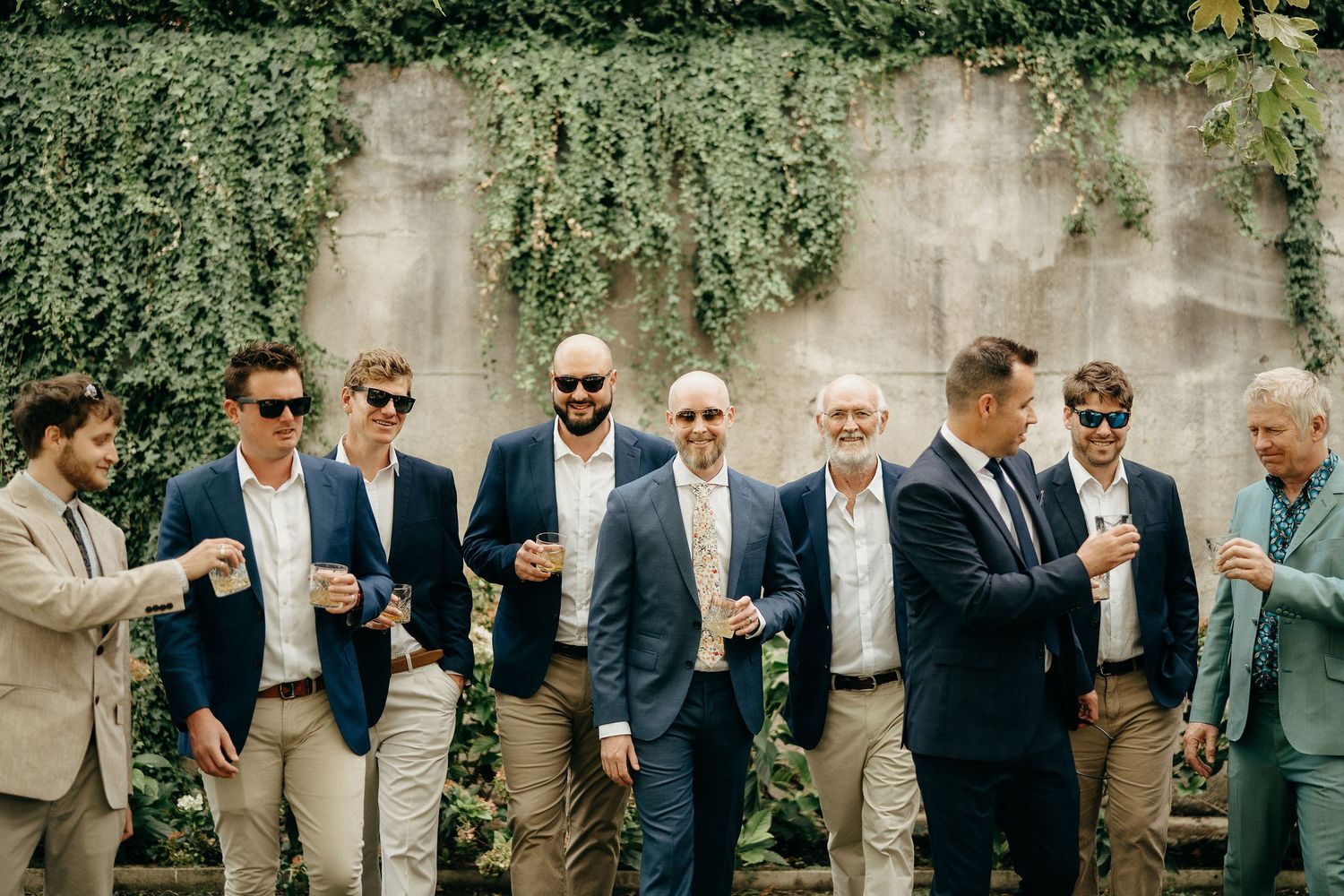 A group of well-dressed men in suits and sunglasses stand together against an ivy-covered wall.