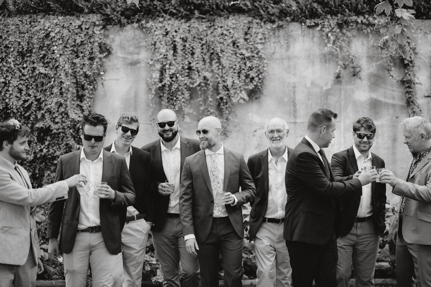 A black and white photograph of a group in suits sharing drinks and laughing together at an outdoor gathering.