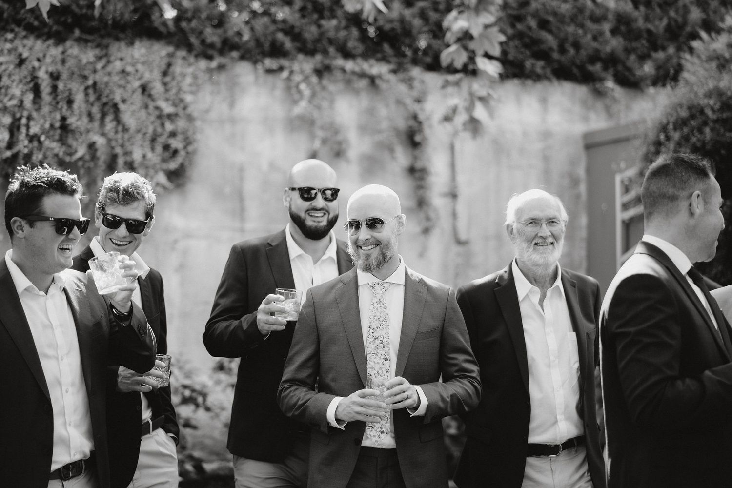 A black and white photograph shows a group of formally dressed men in suits and sunglasses at an outdoor gathering.