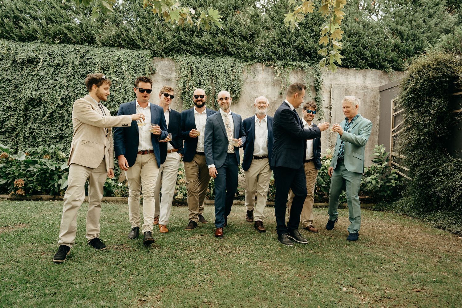 A group in suits and sunglasses sharing casual conversation while standing on a lawn outside.