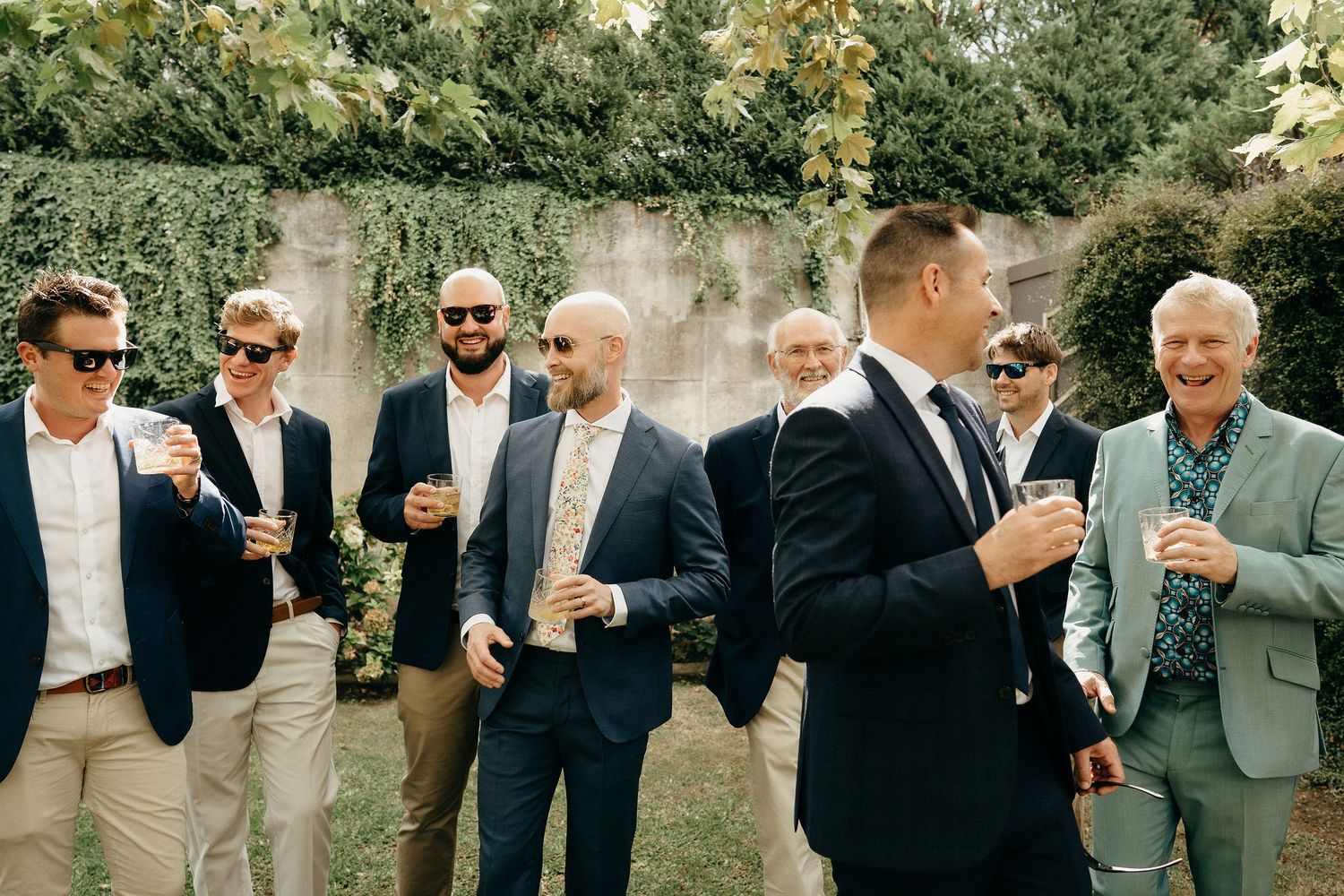 A group of formally dressed people in suits and sunglasses gather outdoors for drinks during a social event.