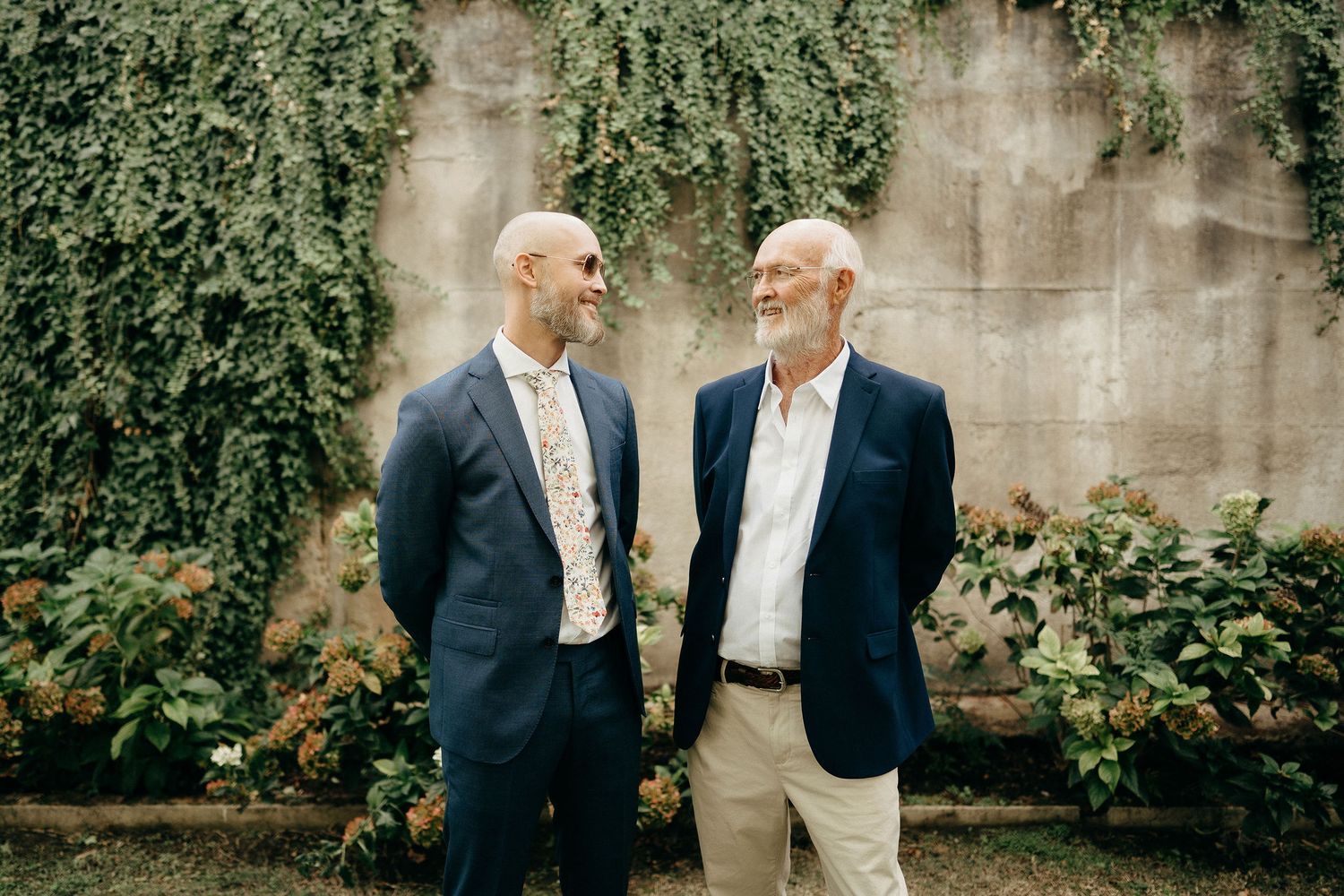 Two men in business casual attire share a candid moment outdoors against a wall with climbing vines.