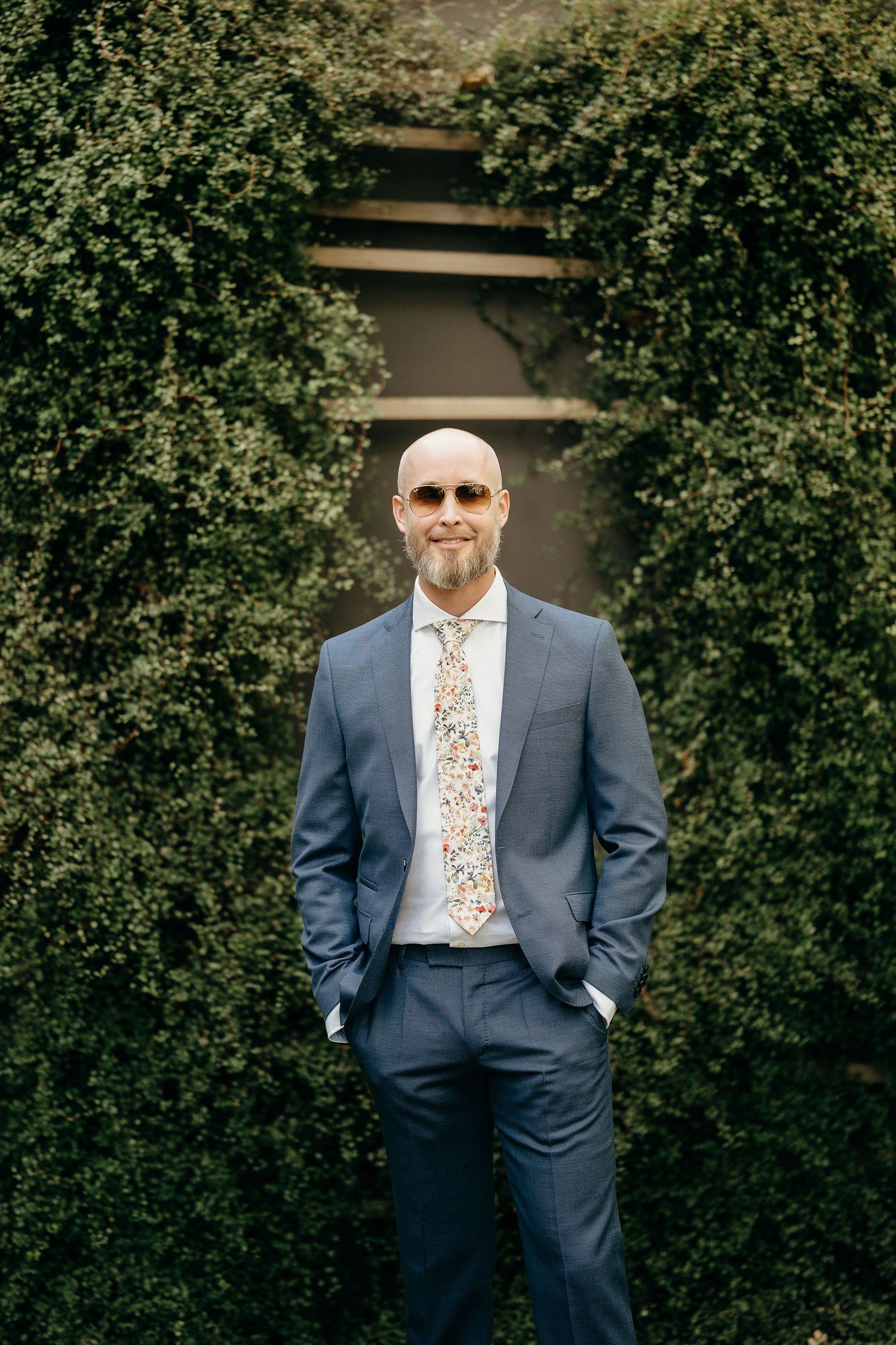 Formal suited figure stands confidently against green hedge wall with steps in background.