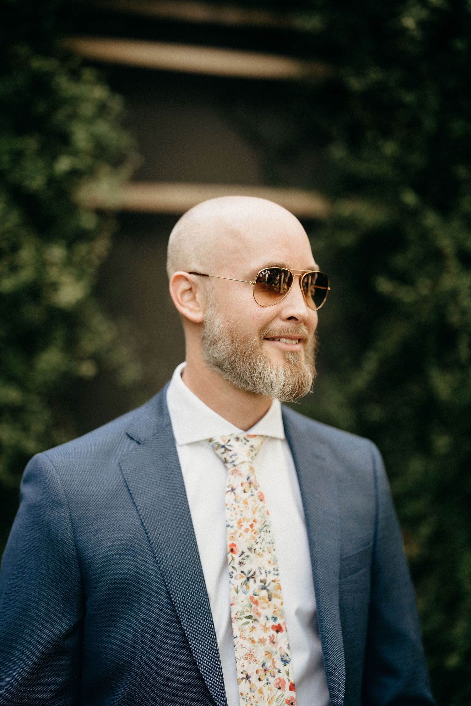 Smiling bald man with grey beard wearing navy suit, floral tie and aviator sunglasses outdoors.
