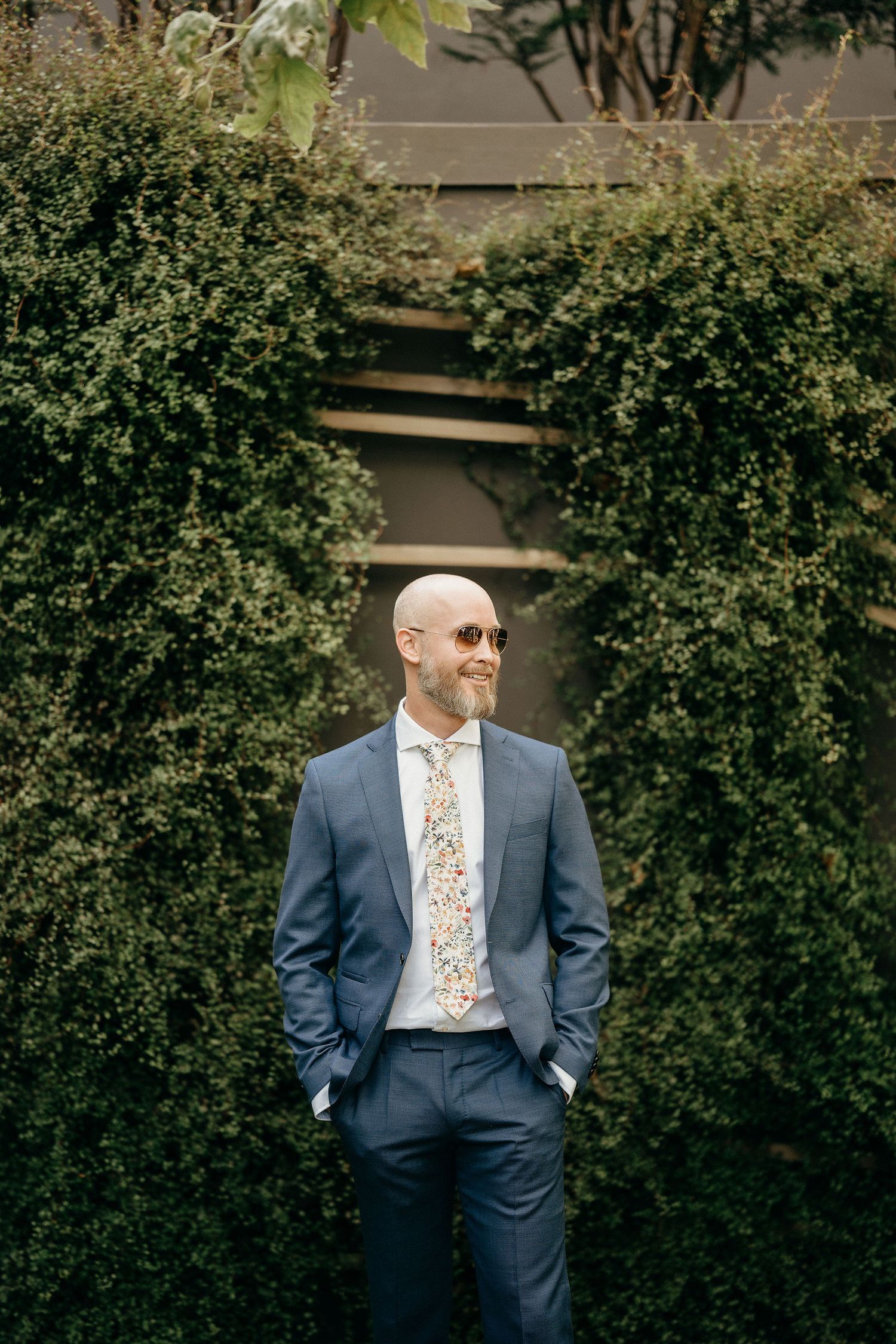 A well-dressed person in a blue suit and floral tie stands against lush green hedges on a sunny day.