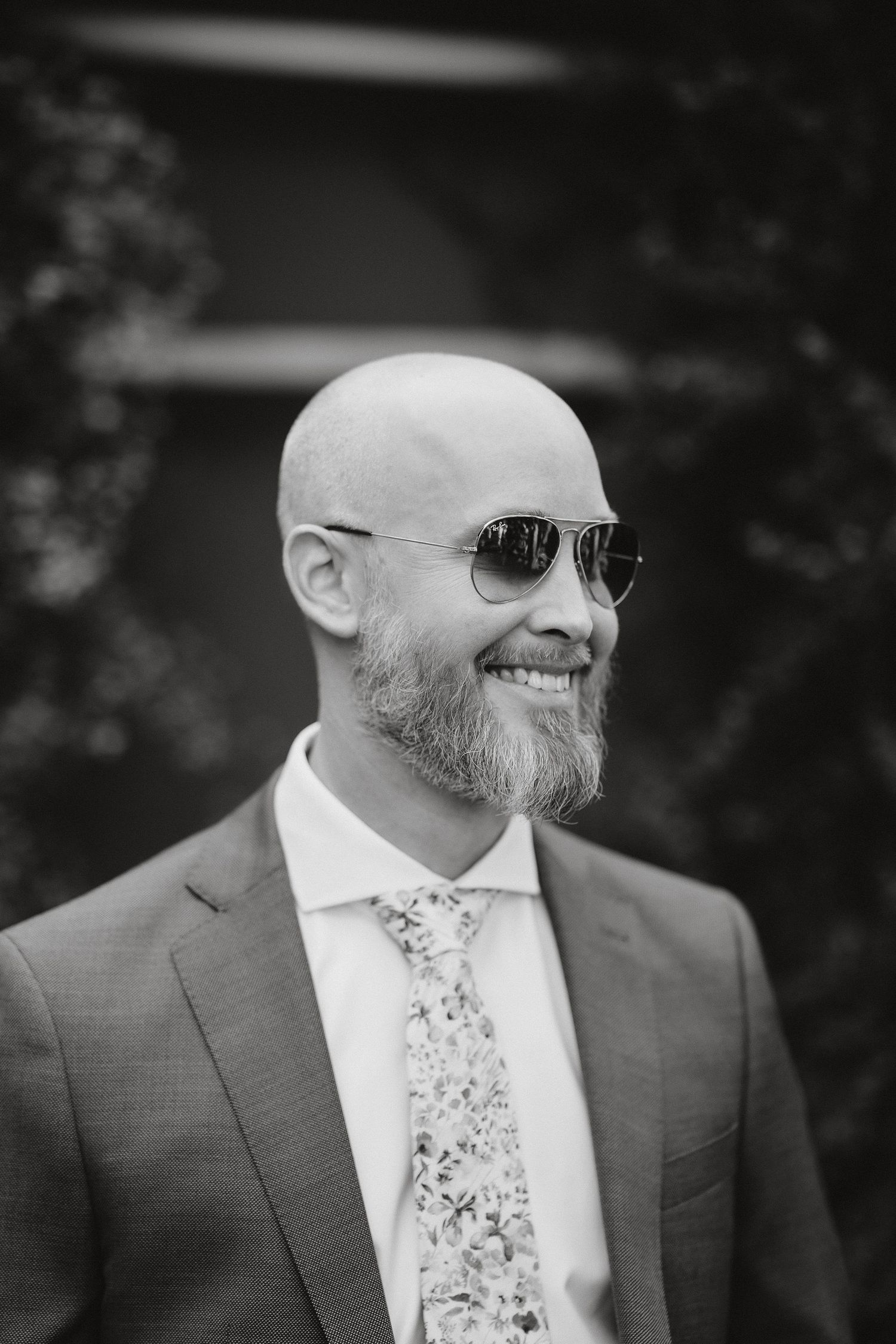 Black and white portrait of a smiling person wearing sunglasses, a floral tie and suit with a neatly trimmed beard.