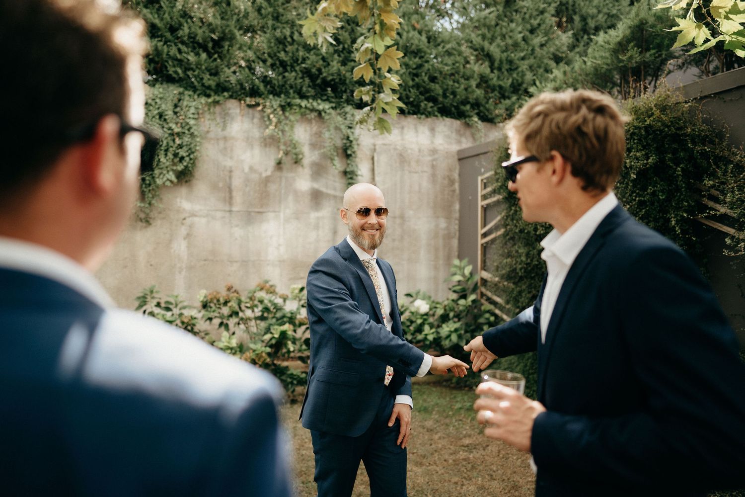 Three people wearing suits having a casual conversation and shaking hands in a garden setting outdoors.