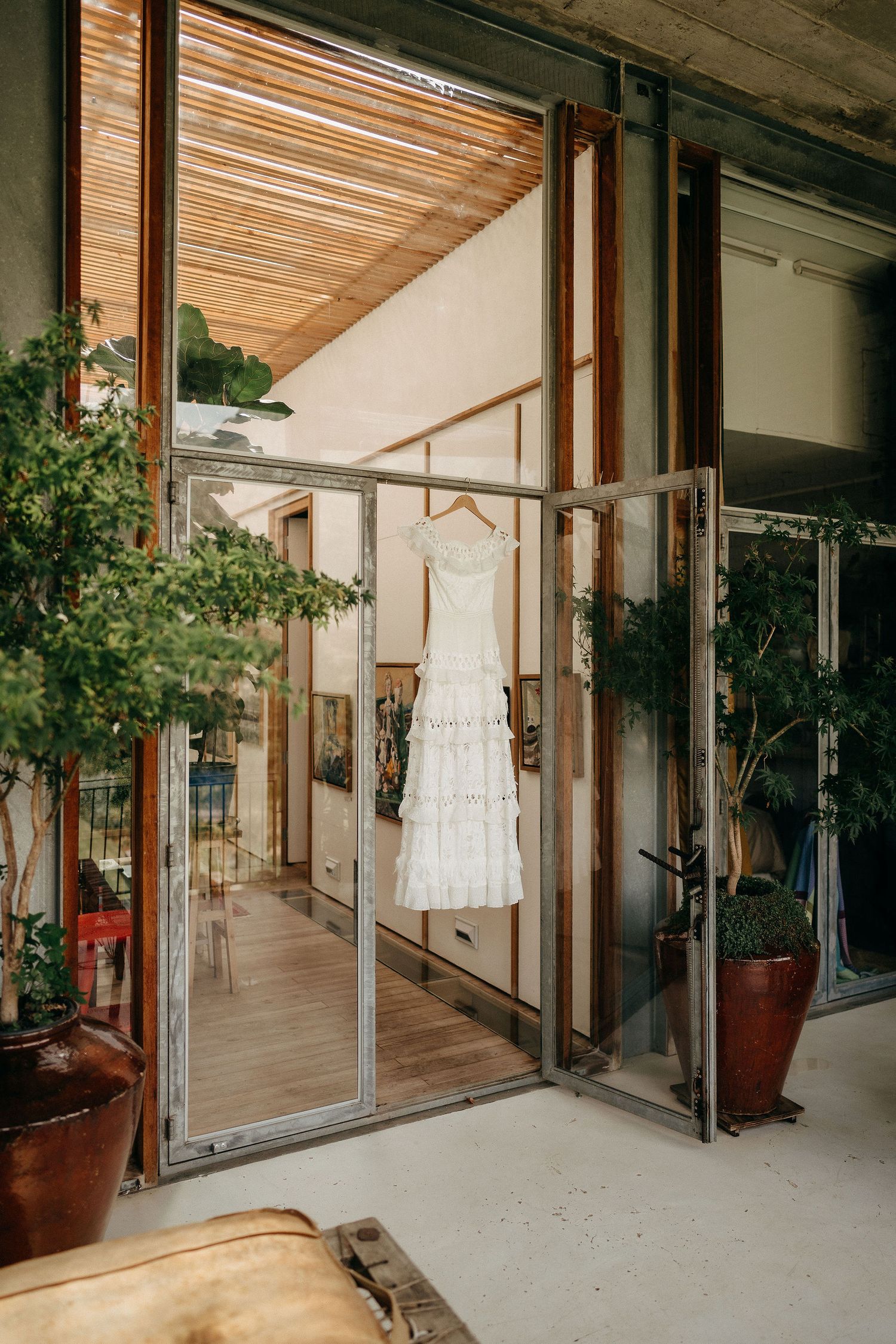 A white lace wedding dress hangs in a boutique storefront with wood accents, glass doors and potted plants.