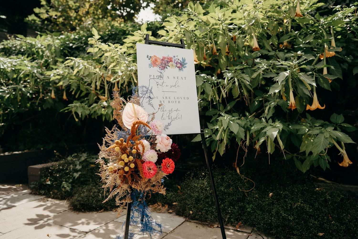 A decorative welcome sign stands outdoors with autumn-colored floral arrangements against lush green foliage.
