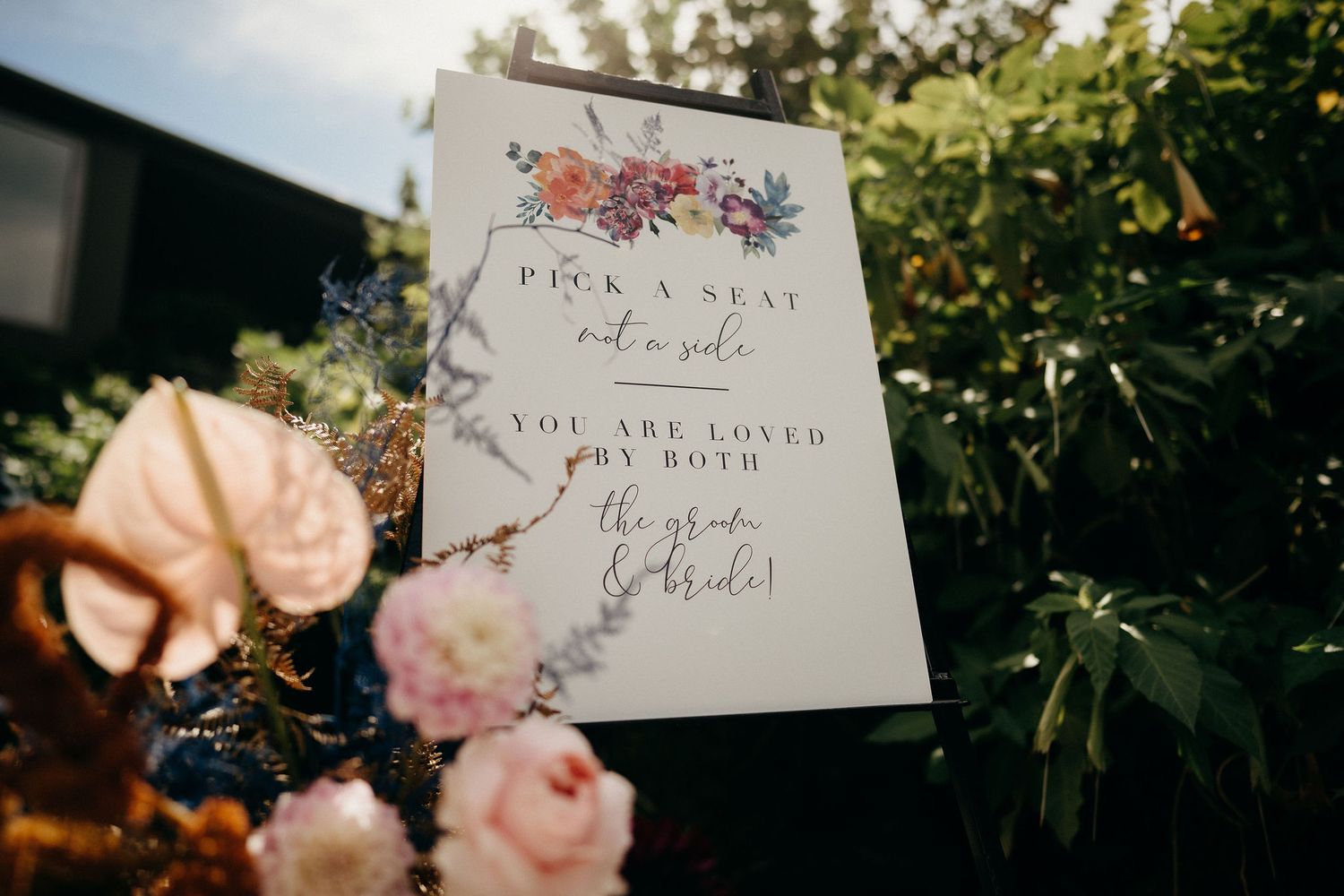 A wedding invitation card with floral designs sits among pink roses in outdoor garden lighting.