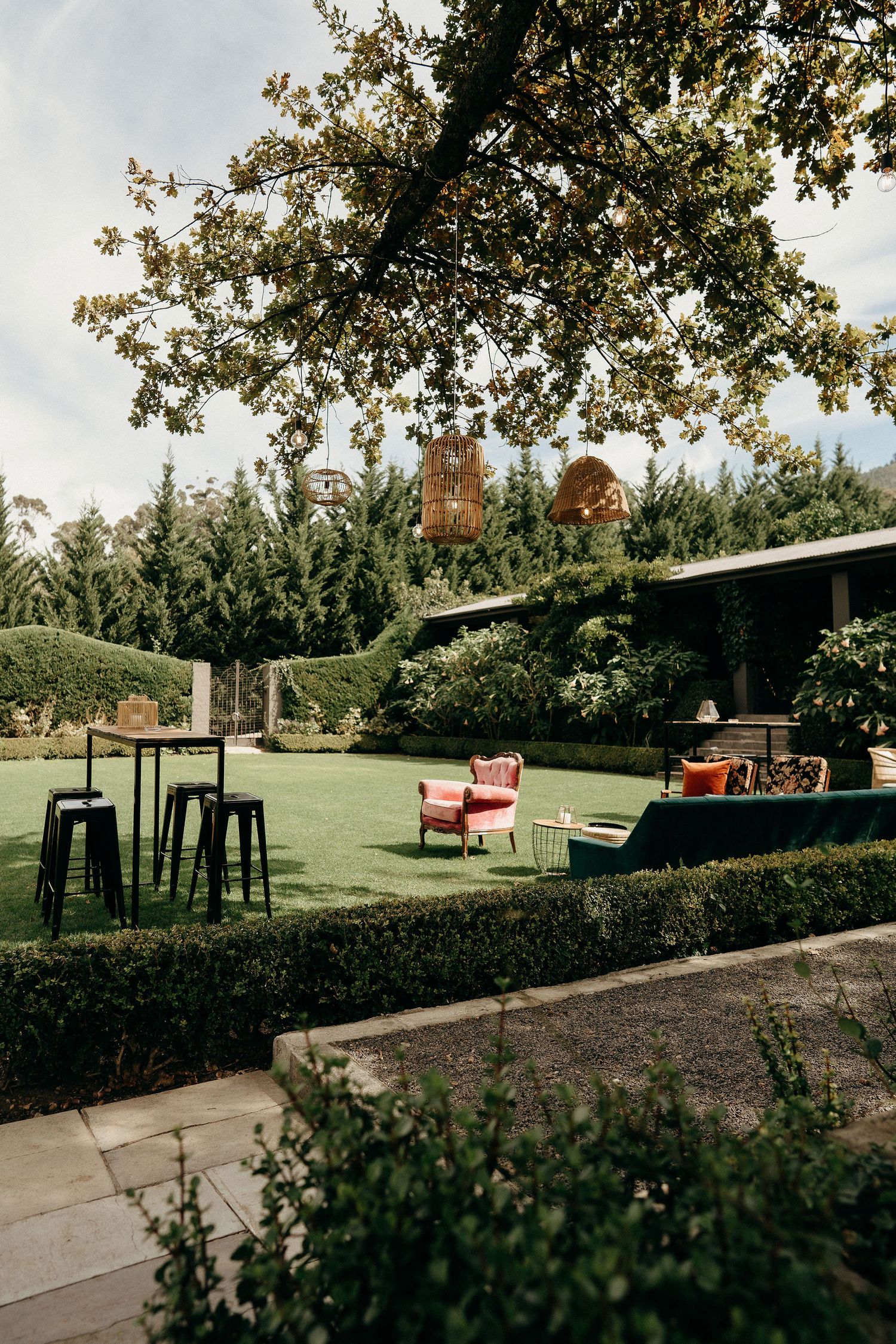 Outdoor garden space with tall bar tables, red accent chairs, and lush greenery under a sprawling tree canopy.