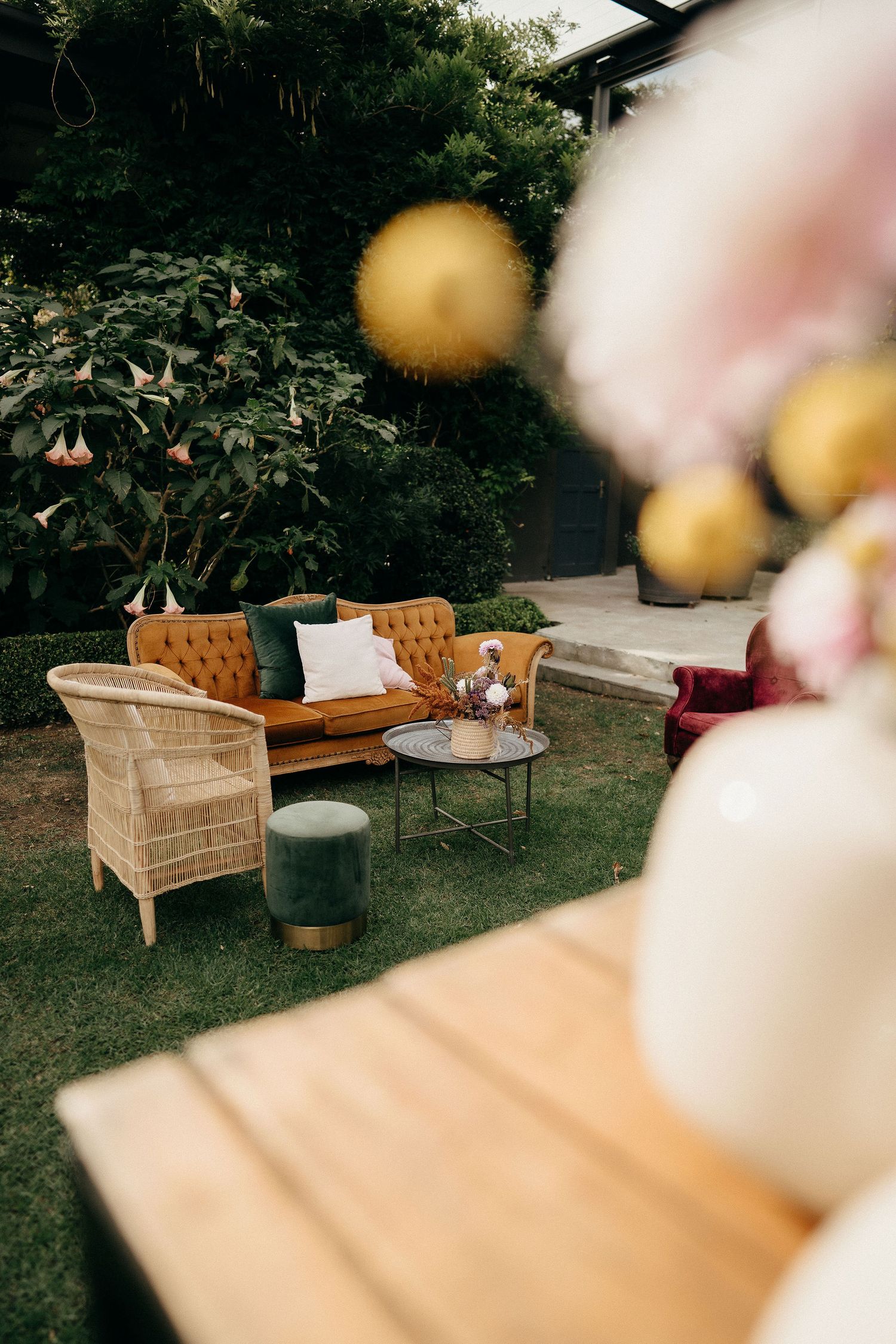 A cozy outdoor seating area with a brown leather couch, wicker chair, and decorative gold lanterns in a garden setting.