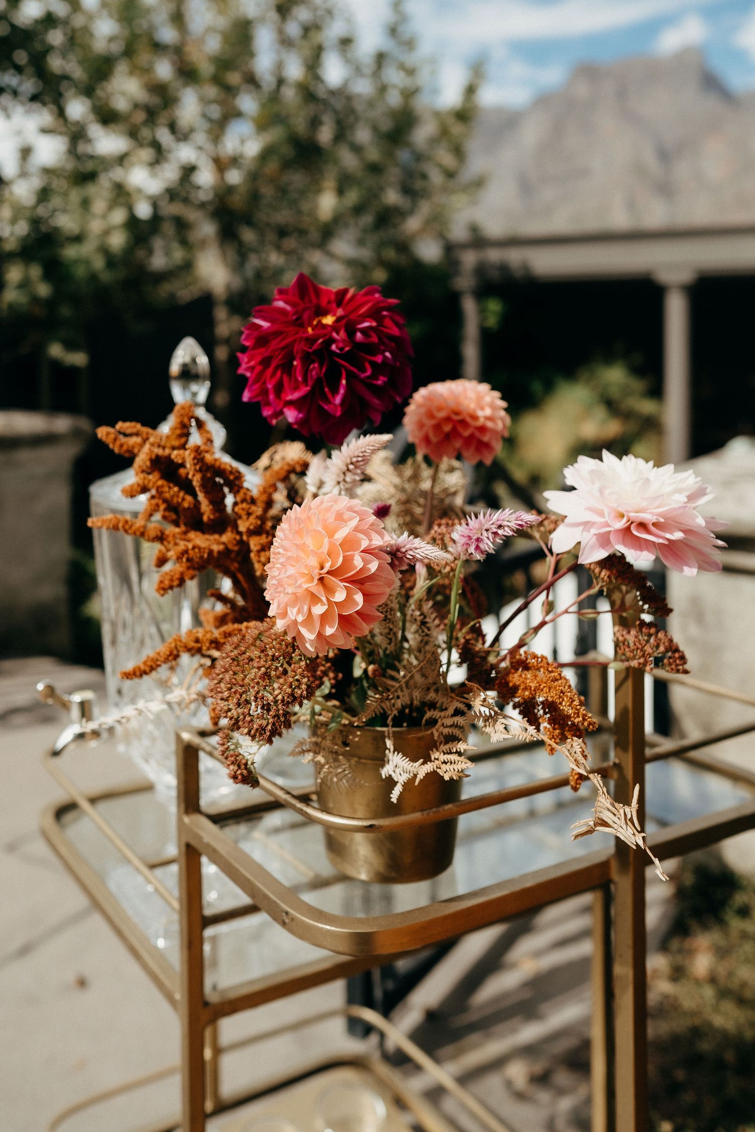 Beautiful floral arrangement with red and pink dahlias in gold vase on vintage metal bar cart outdoors.