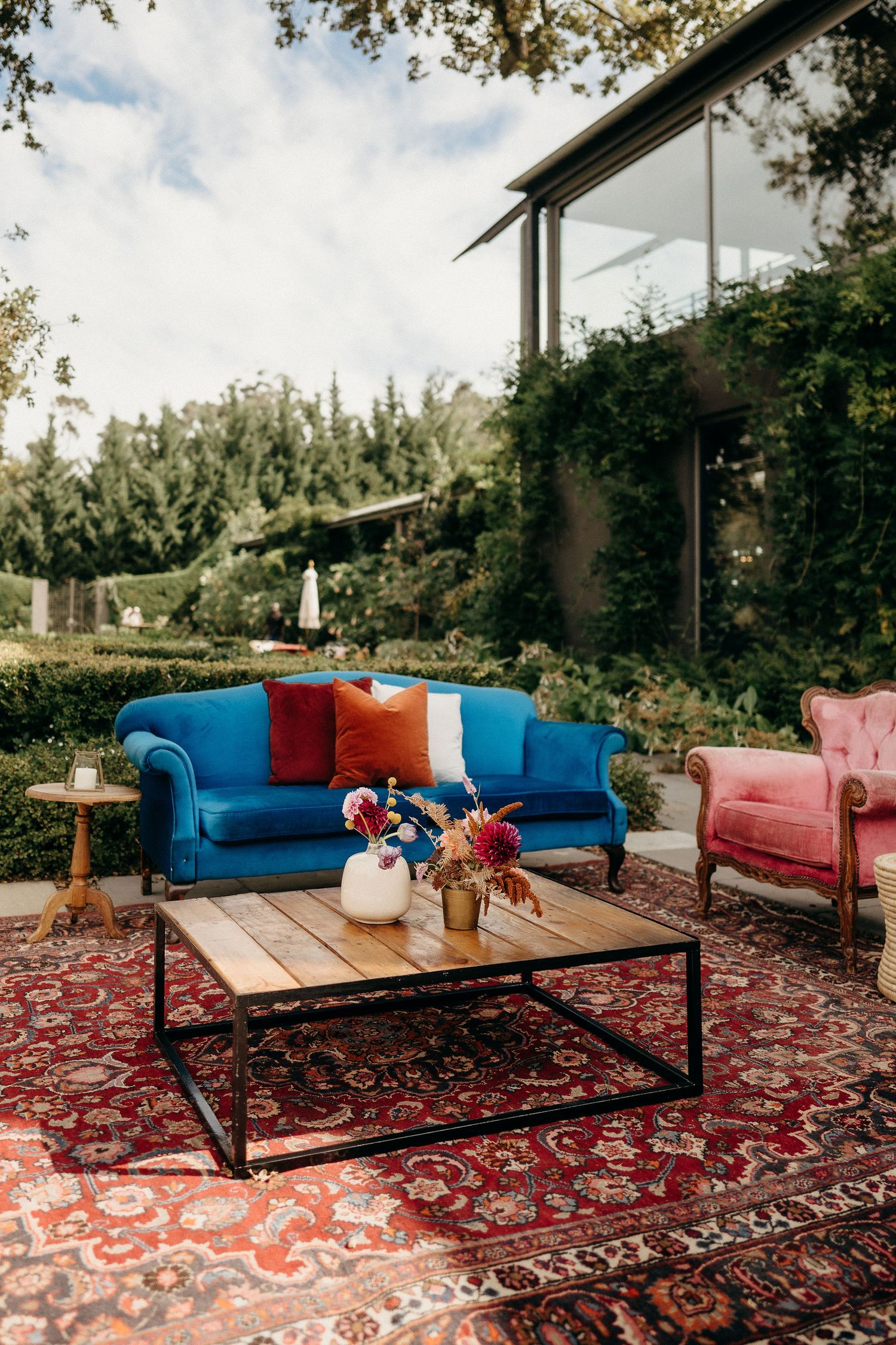 A cozy outdoor lounge area features a blue velvet sofa, pink armchair, and wooden coffee table on a vintage oriental rug.