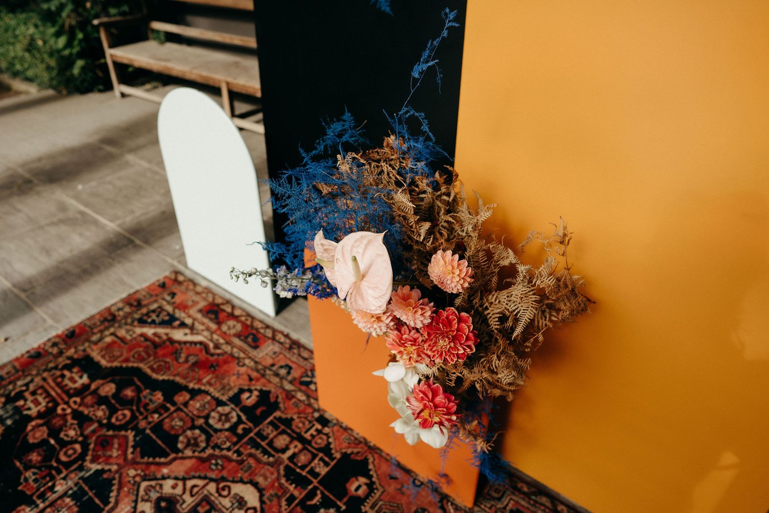 A decorative corner display with a Persian rug, surfboard, and dried floral arrangement against yellow and blue walls.