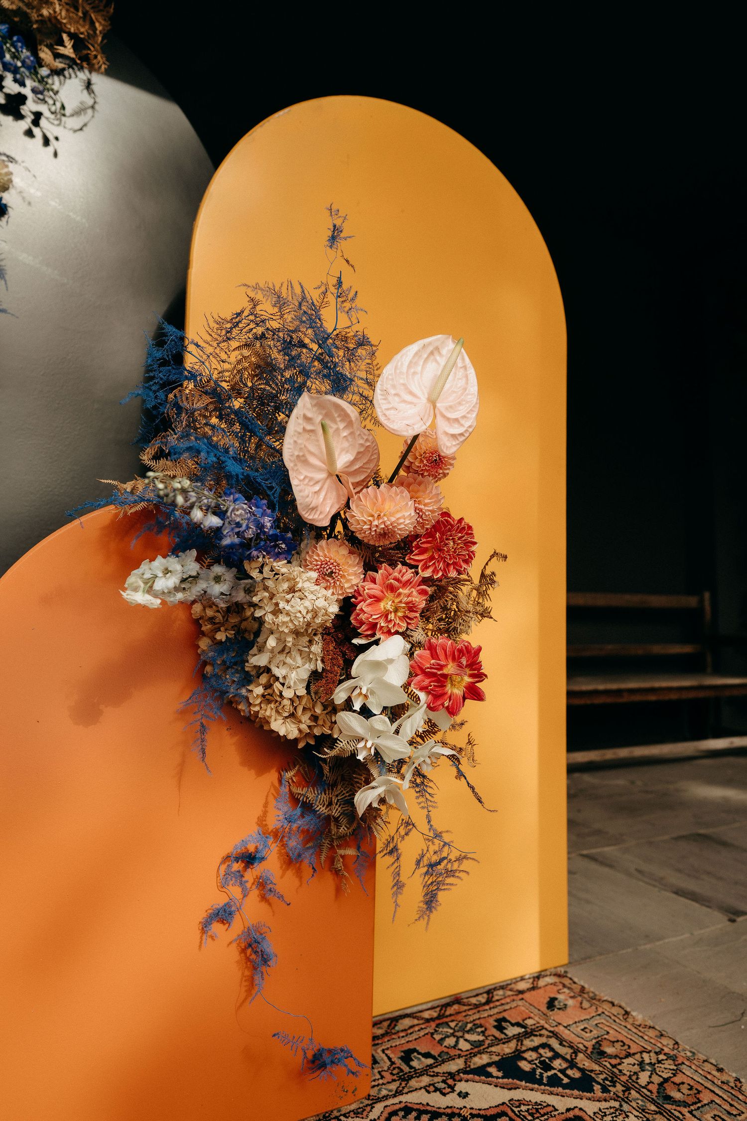 A floral arrangement with dried flowers in blue, red and white blooms against an orange arched wall with an ornate rug below.