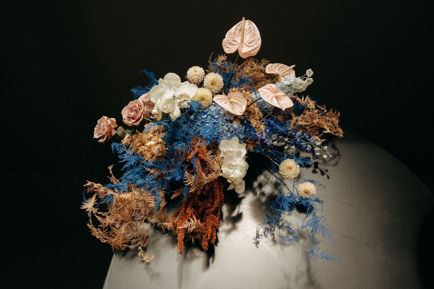 A romantic wedding floral arrangement with dried blue delphinium, white roses and copper-toned flowers against dark backdrop.