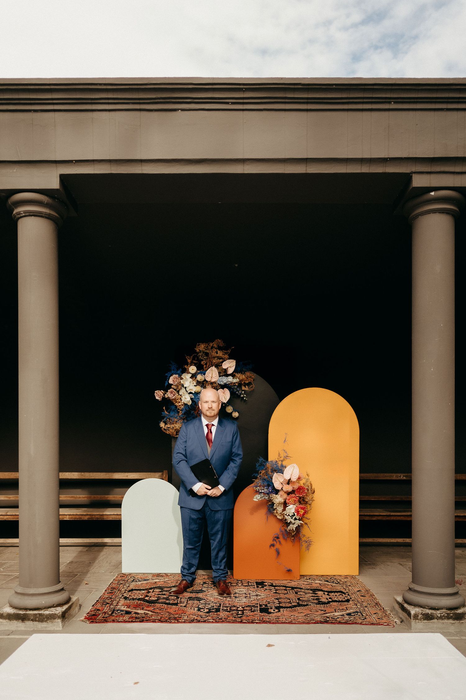 A person in a navy suit stands between ornate floral arrangements and decorative arches on a patterned rug.