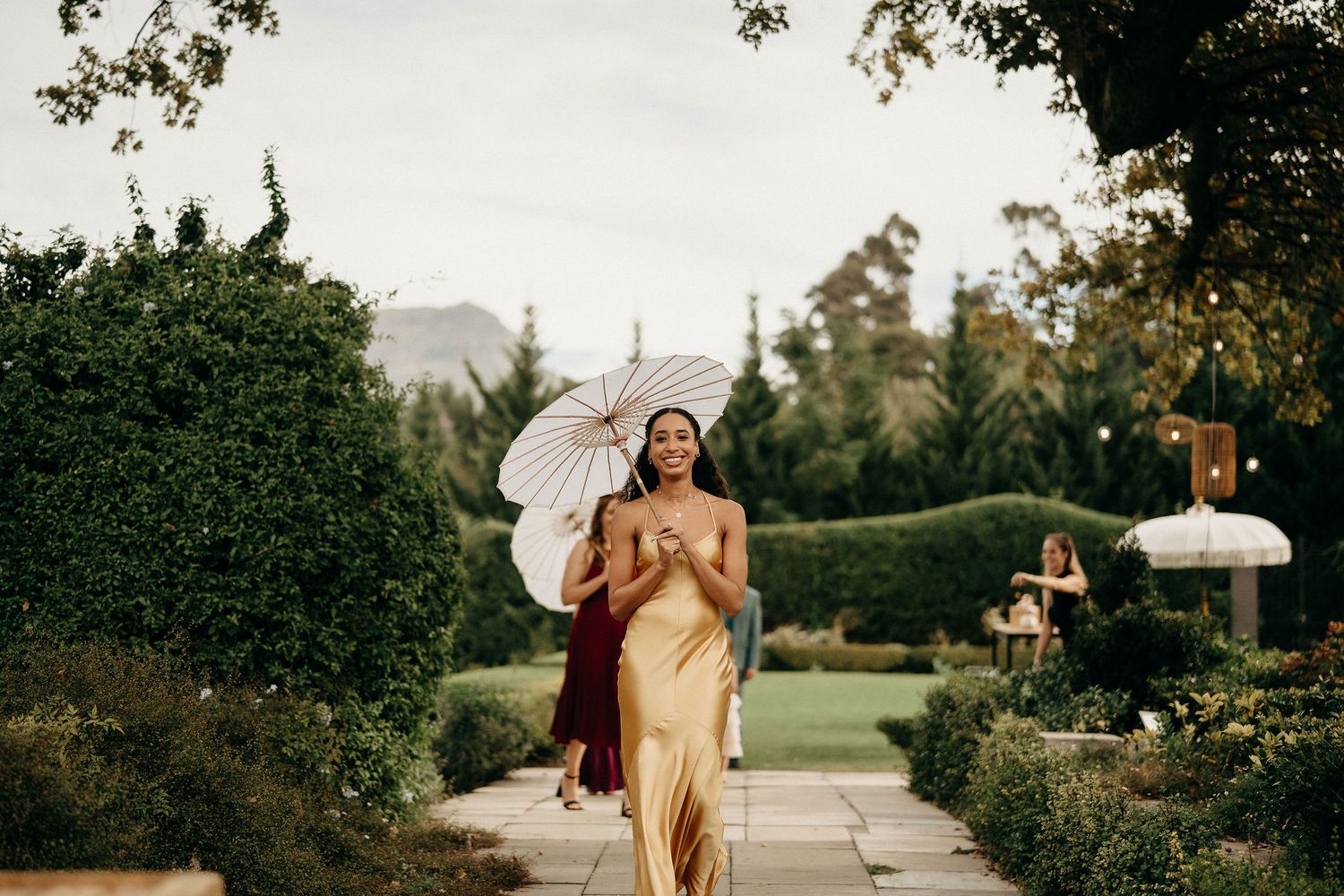 A person in a gold silk dress walks along a garden path holding a white parasol umbrella on a sunny afternoon.