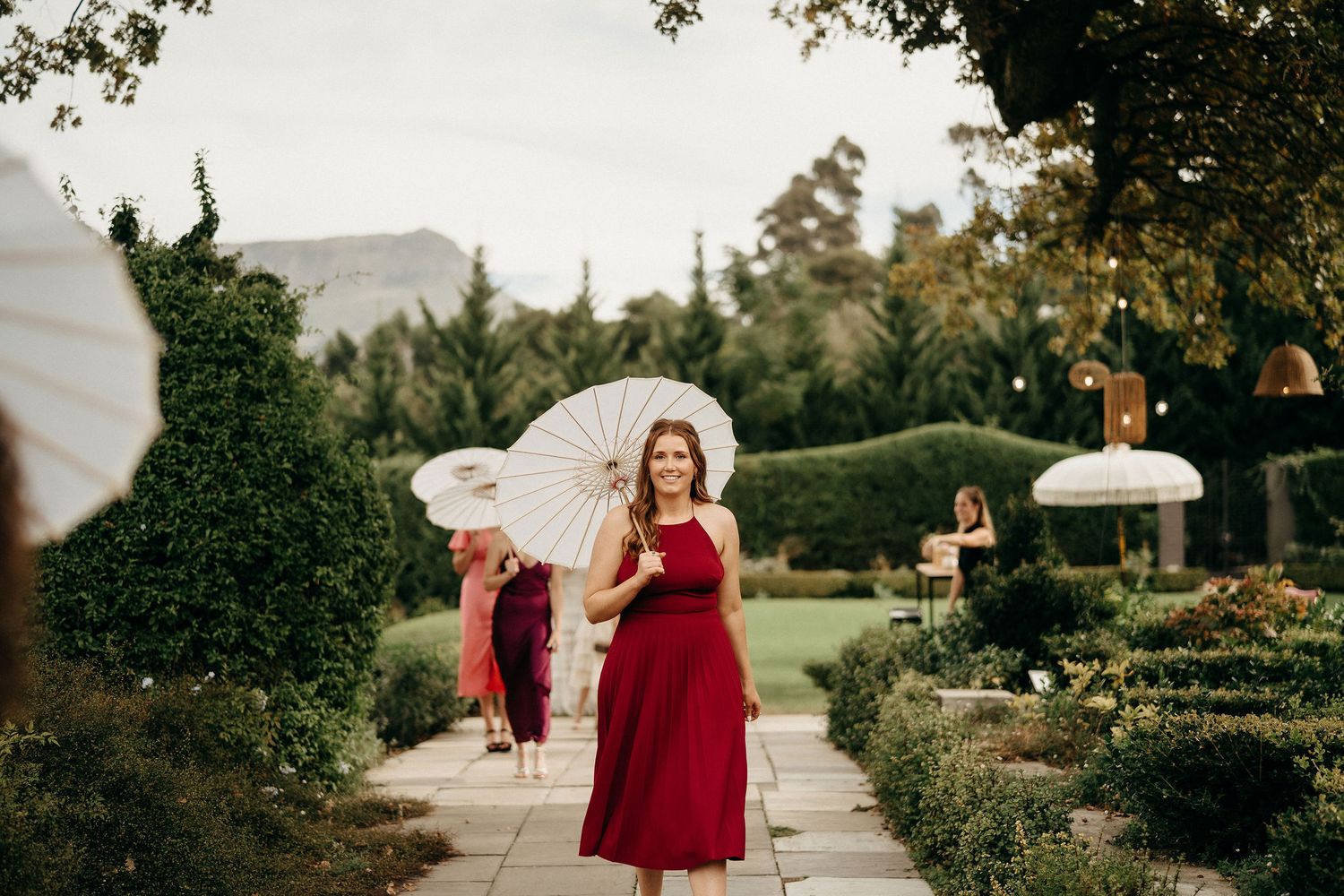 People in elegant attire gather in a lush garden setting with white parasols and wine glasses on a summer afternoon.