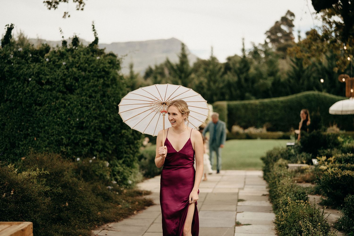 A person in a burgundy satin dress holds a white parasol while walking through a garden path at dusk.