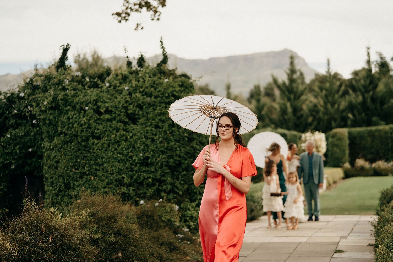 Someone in a vibrant coral dress walking with a white parasol in a garden path with mountains in background.