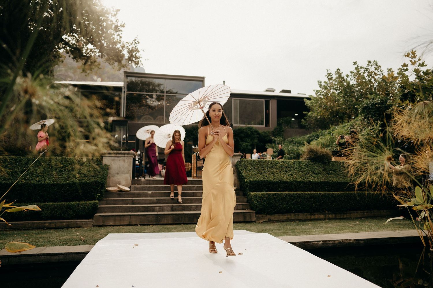 A person in a champagne-colored dress walks along a white aisle during an outdoor celebration at a modern venue.