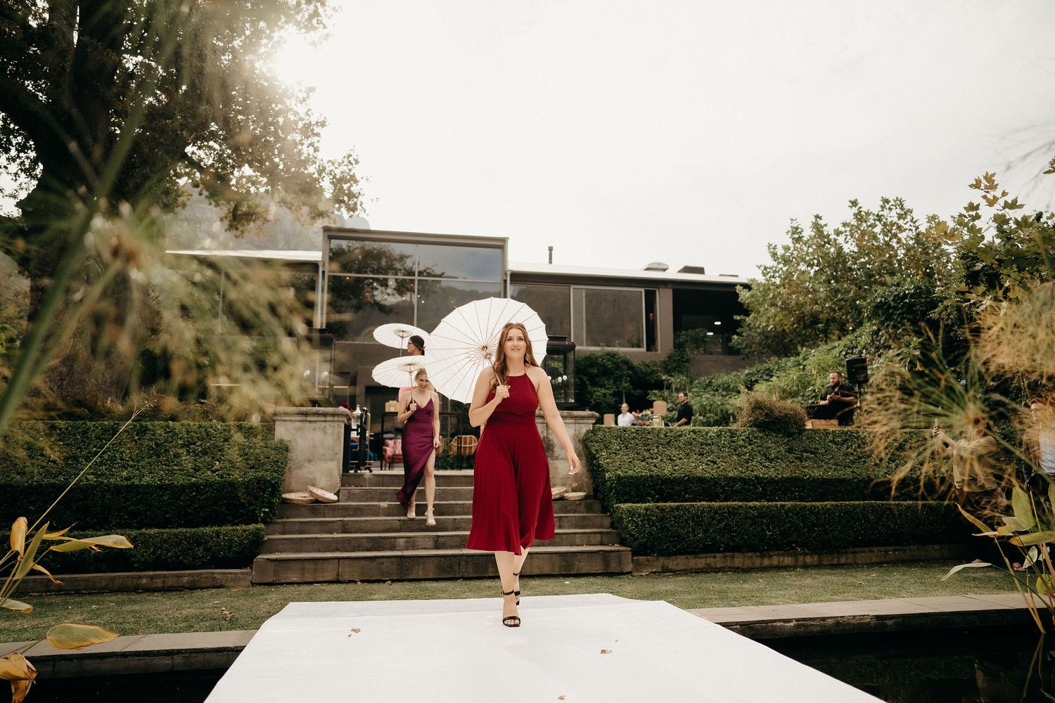 A figure in a flowing red dress walks down outdoor garden steps holding a white parasol on a sunny day.