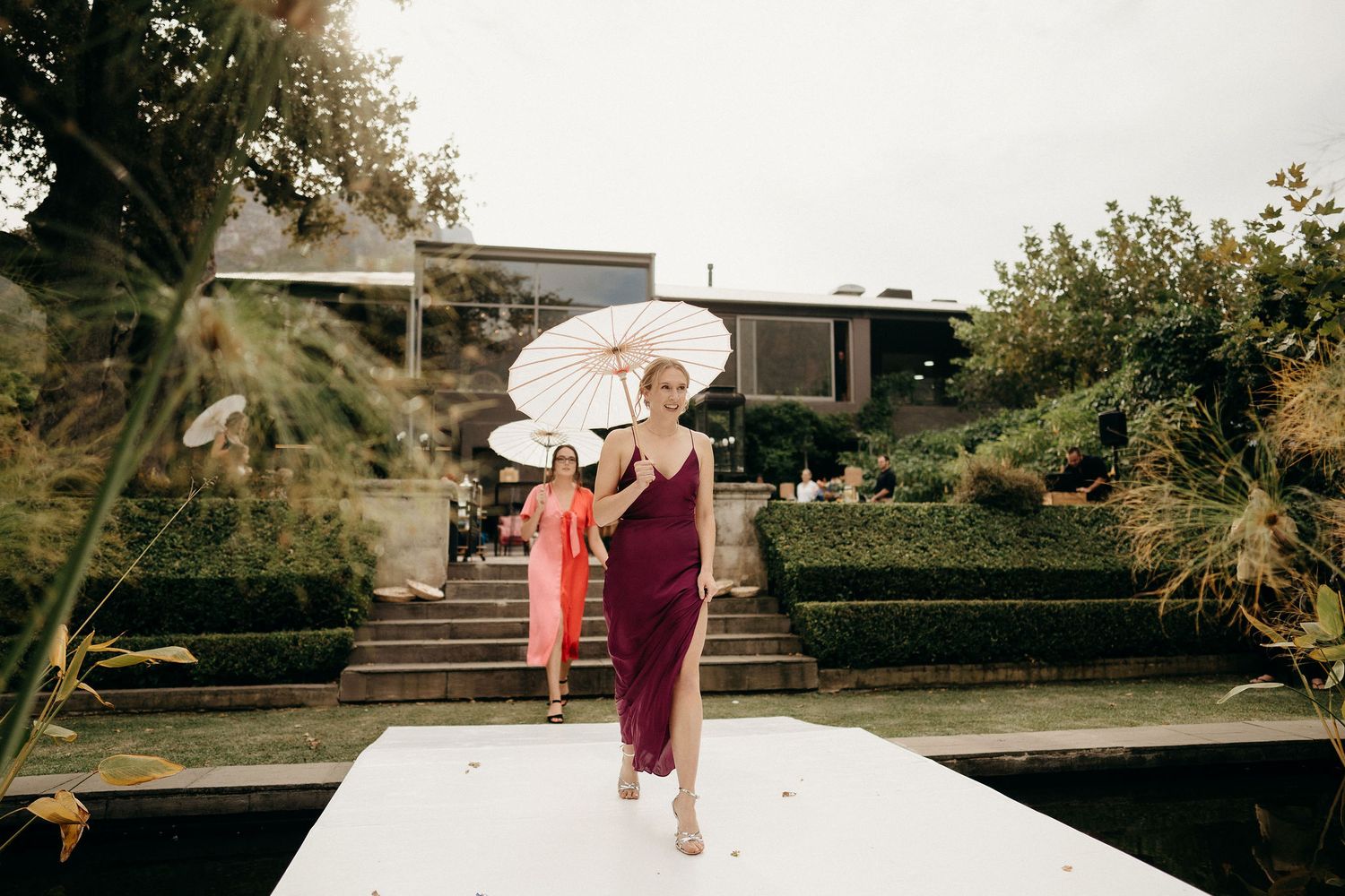 People holding parasols walk on outdoor pathway with garden steps and greenery in the background on a sunny day.