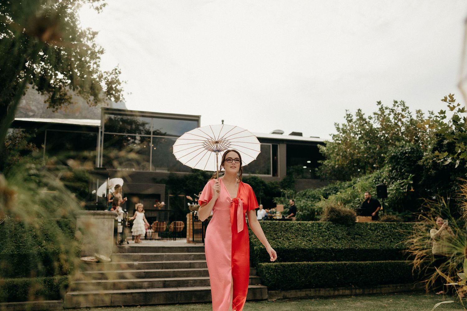 Person in a flowing red dress walks down garden steps while holding a white parasol umbrella on a cloudy day.