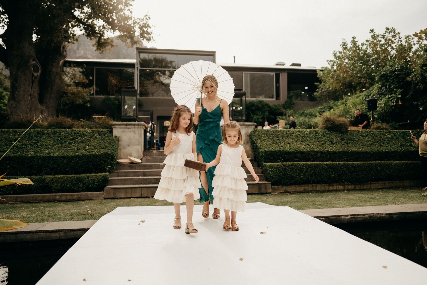 A woman in a green dress stands with two young children in white dresses on an outdoor walkway at sunset.