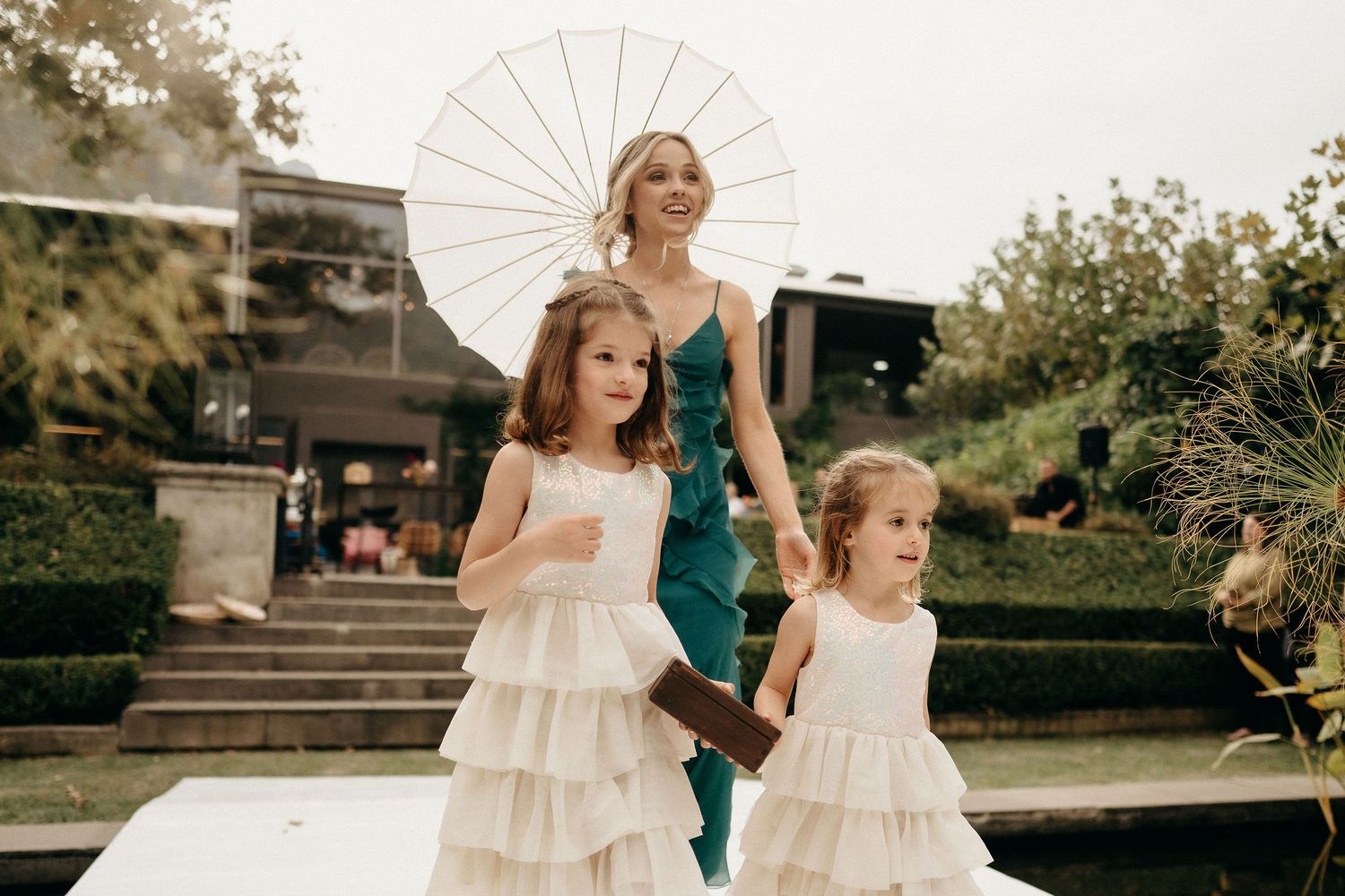 Elegant ladies in green and cream dresses walk outdoors holding a white parasol during a formal event.