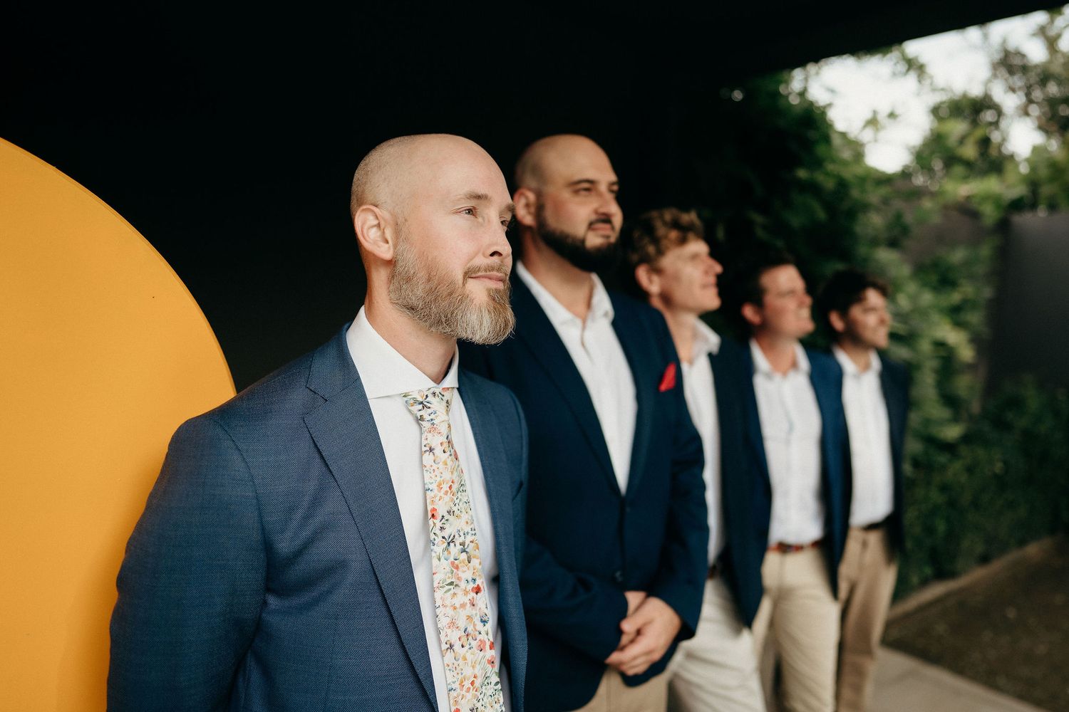 Groomsmen in mixed formal attire lined up outdoors with trees and shrubbery in background.