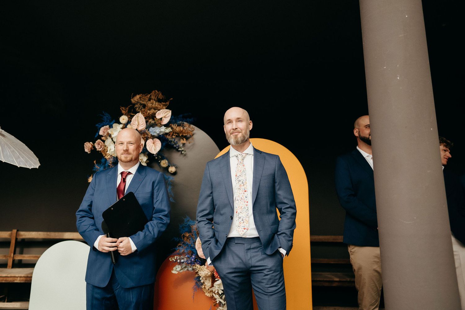 Three well-dressed men stand together in a modern space, with one wearing a navy suit and another in gray suit.