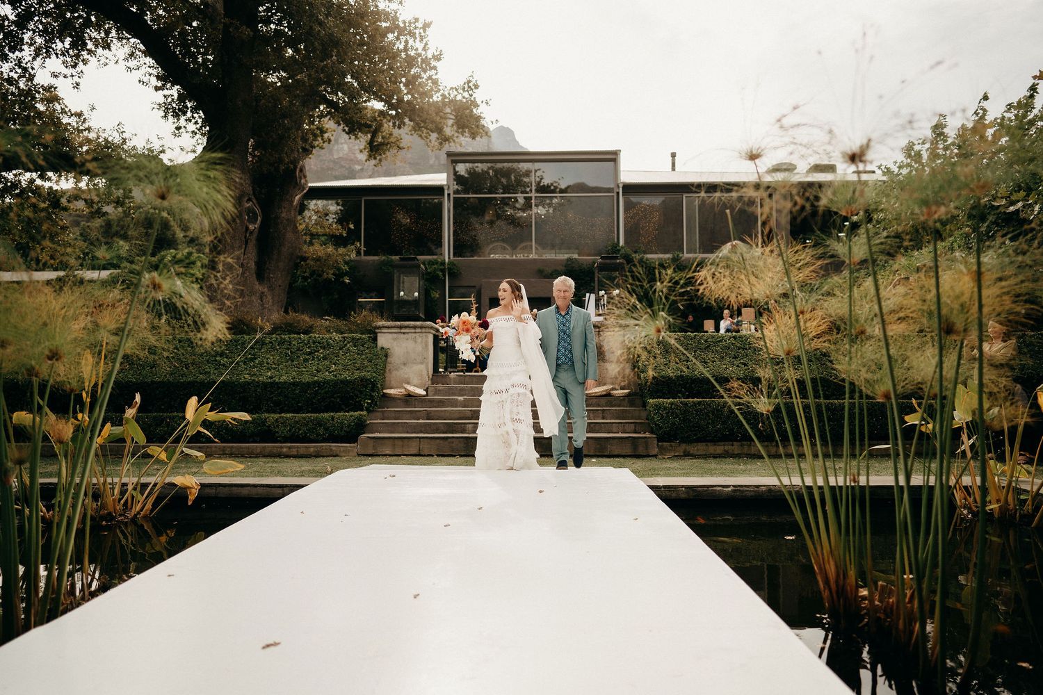 A wedding couple standing on a white walkway surrounded by a pond and lush garden at a modern venue.