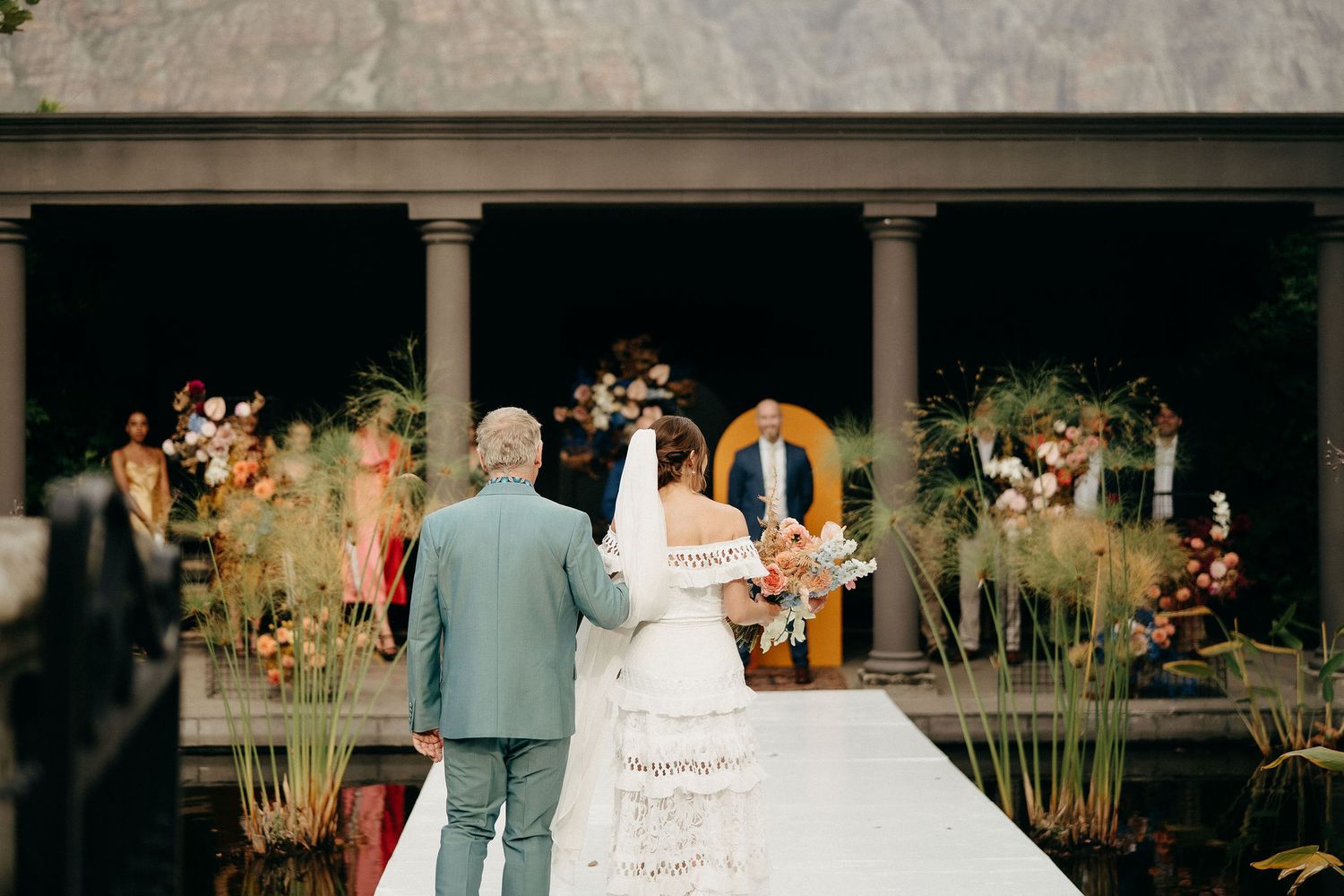 An outdoor wedding ceremony takes place under a columned pavilion surrounded by decorative grasses and flowers on a white aisle.