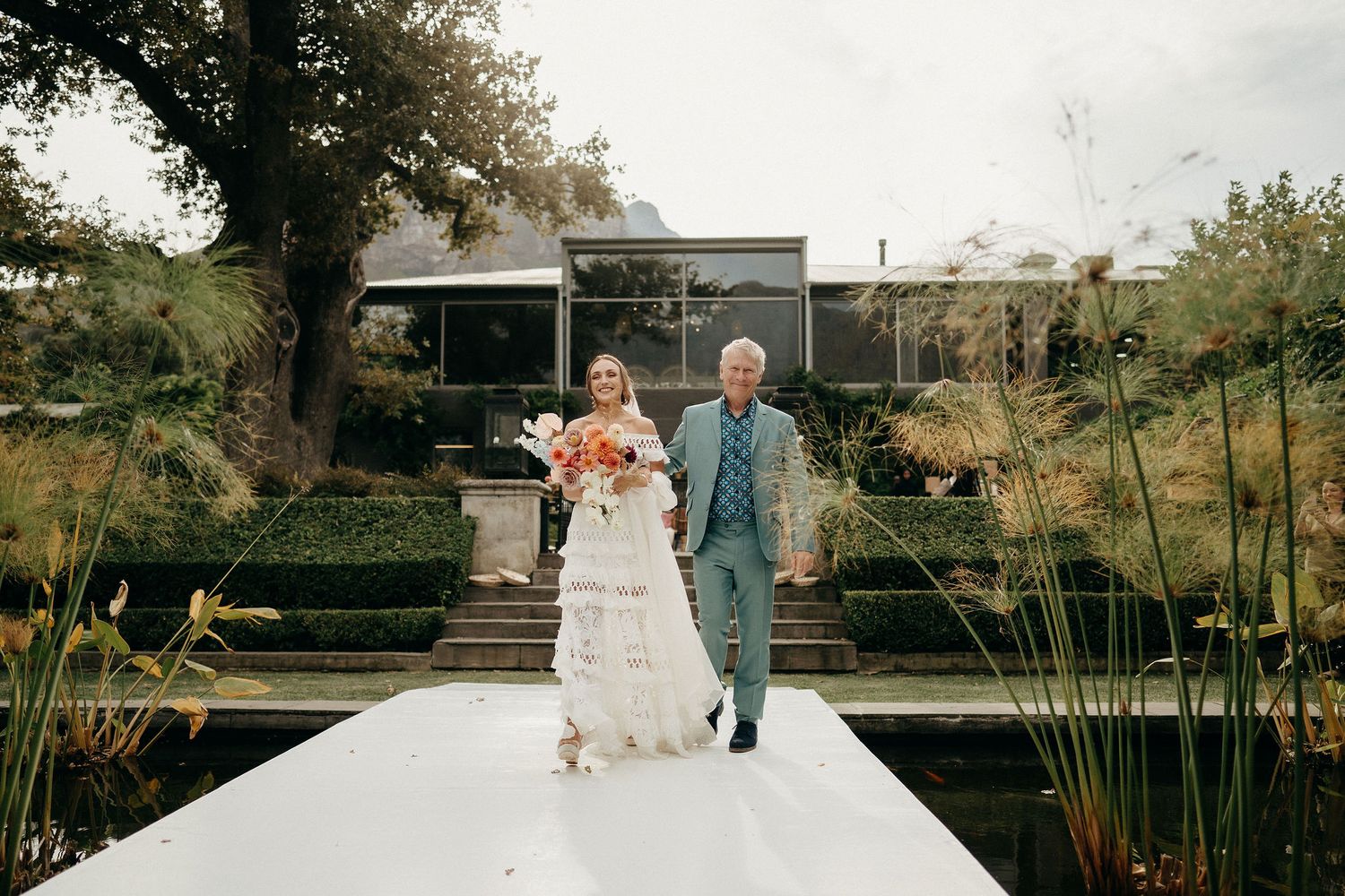 A couple walks down a white aisle at an outdoor wedding venue with a modern house and natural landscaping in the background.