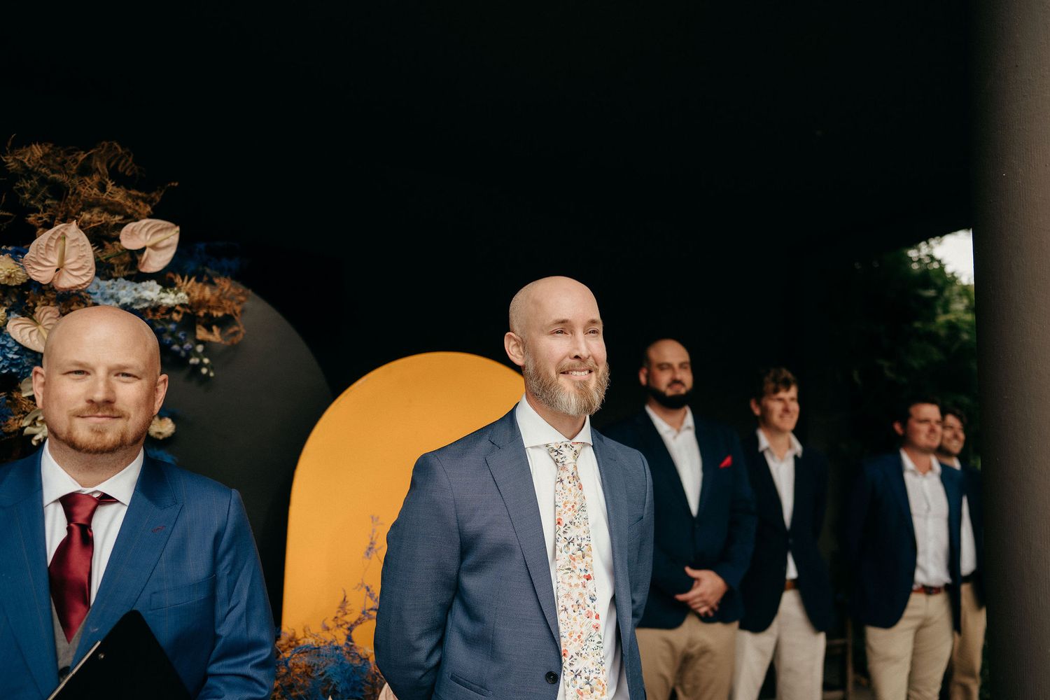 A group of well-dressed groomsmen stand together before a modern backdrop with yellow and black geometric shapes.