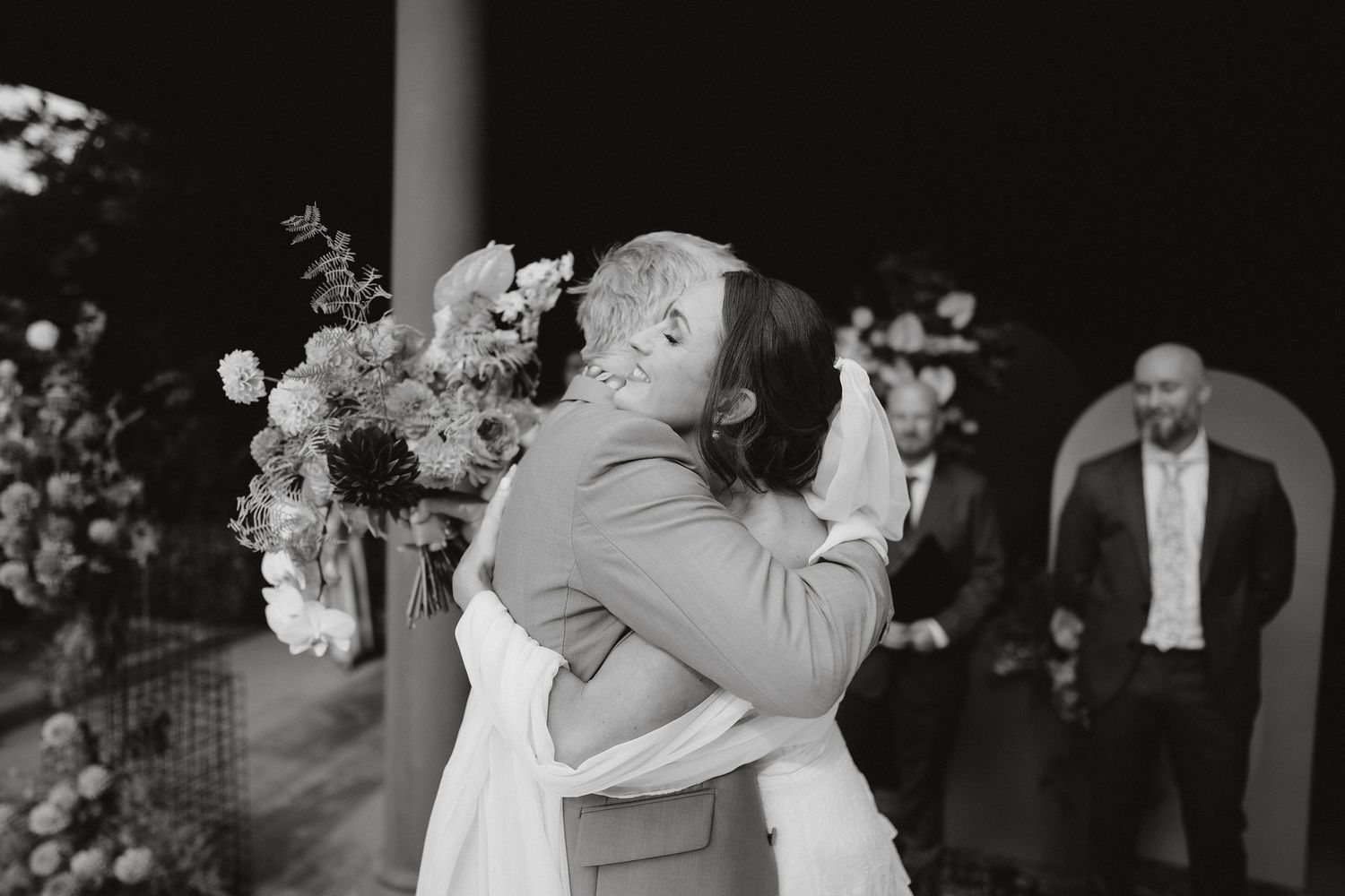A tender embrace between two people at a wedding ceremony with floral decorations in black and white photography.