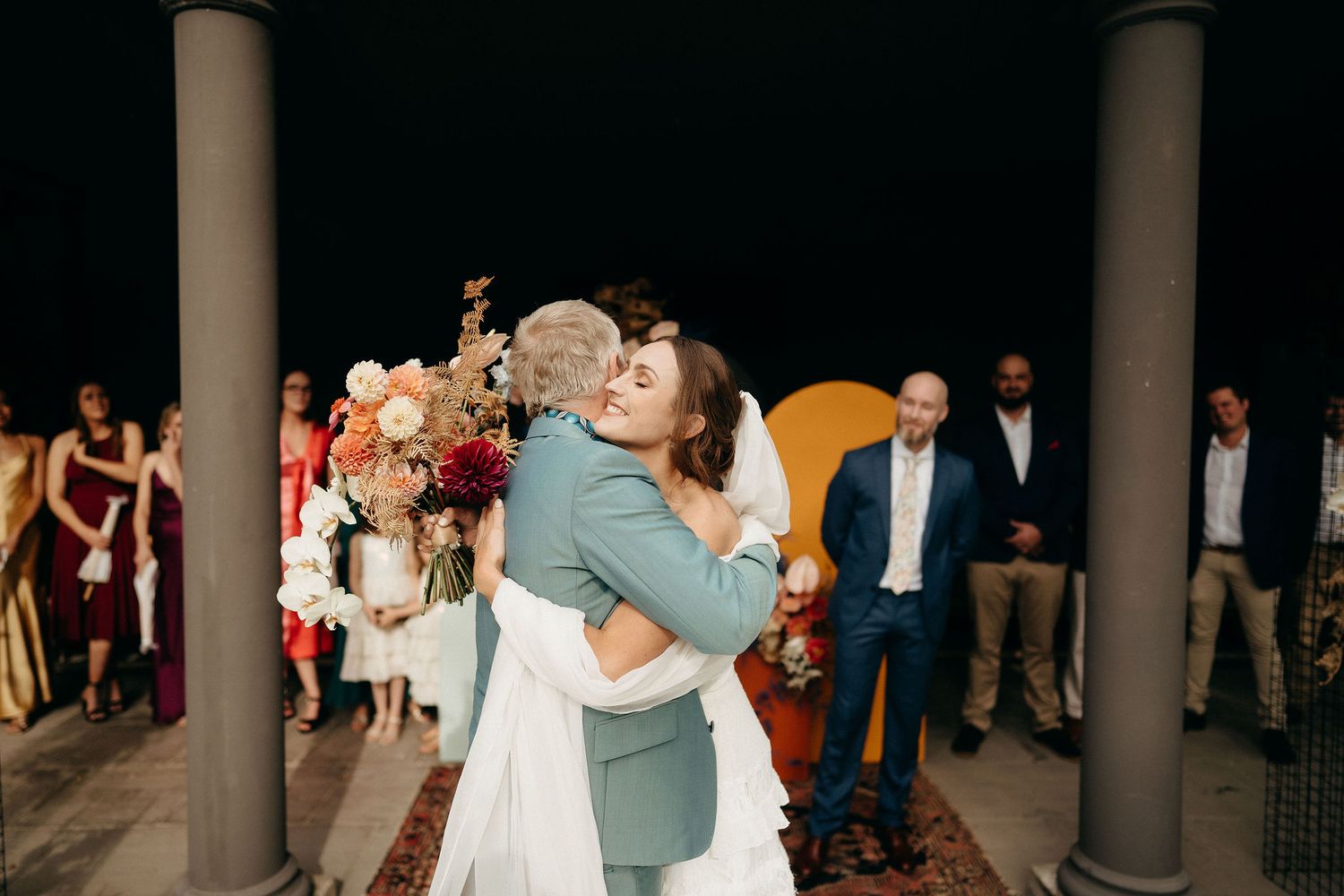 A couple embraces in a tender moment during their wedding celebration surrounded by guests on a covered patio.