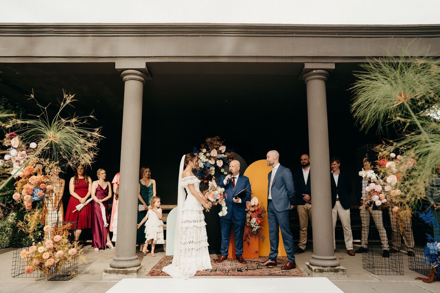 A wedding ceremony takes place under an outdoor pavilion with elegant columns and floral arrangements.