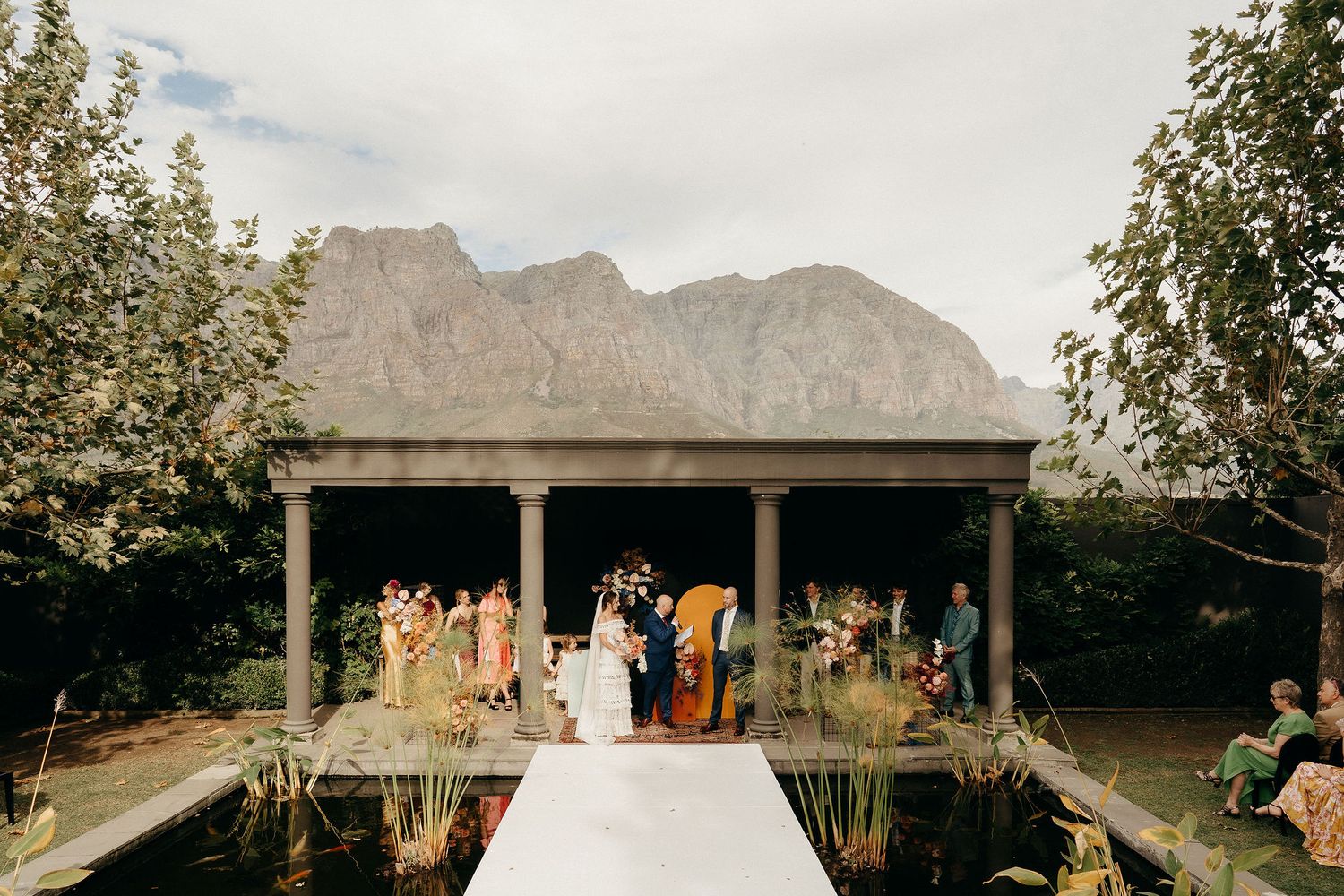 A wedding ceremony takes place at an outdoor pavilion with majestic mountain peaks as the backdrop.