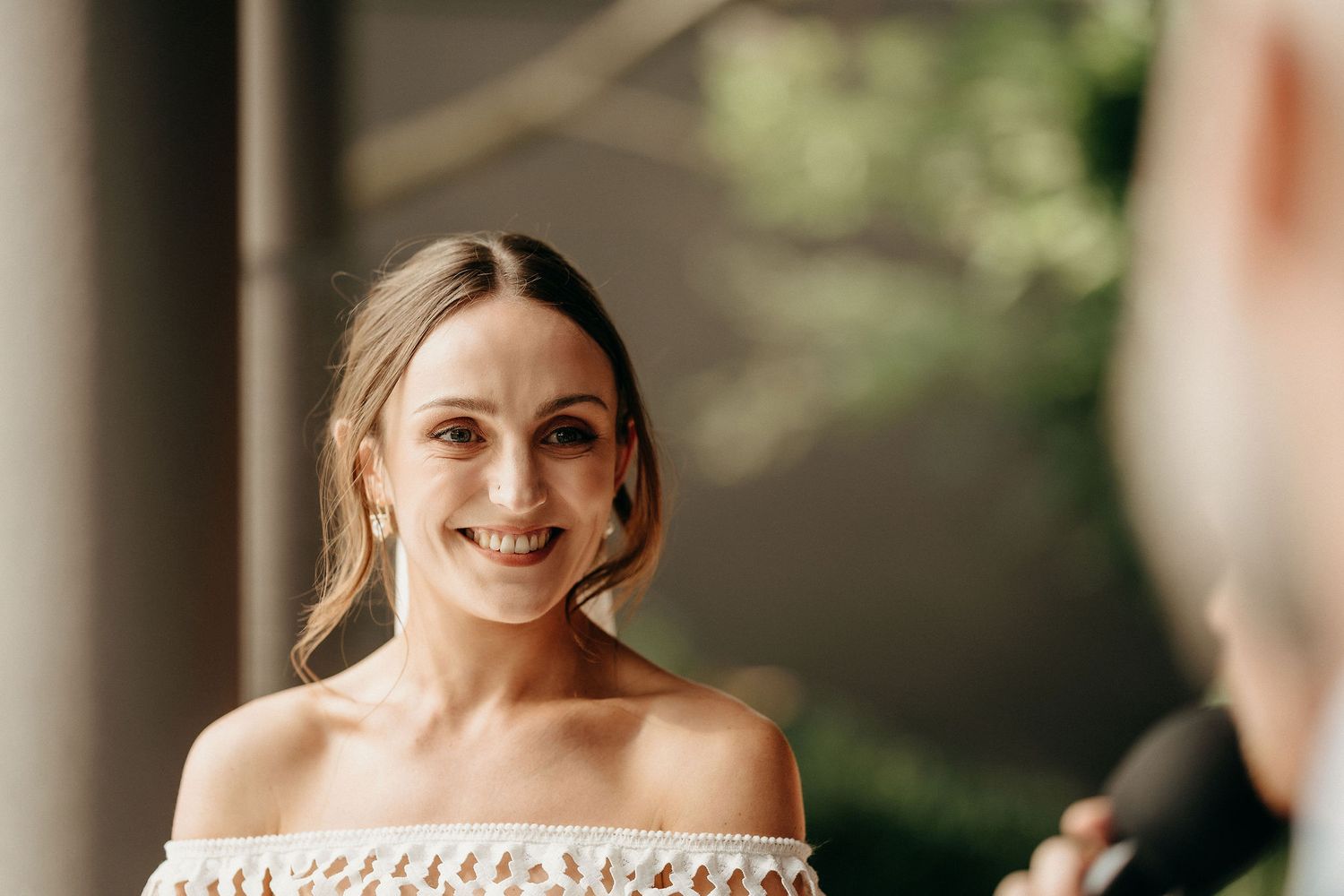 A smiling woman wearing an off-the-shoulder dress poses outdoors in soft natural lighting.