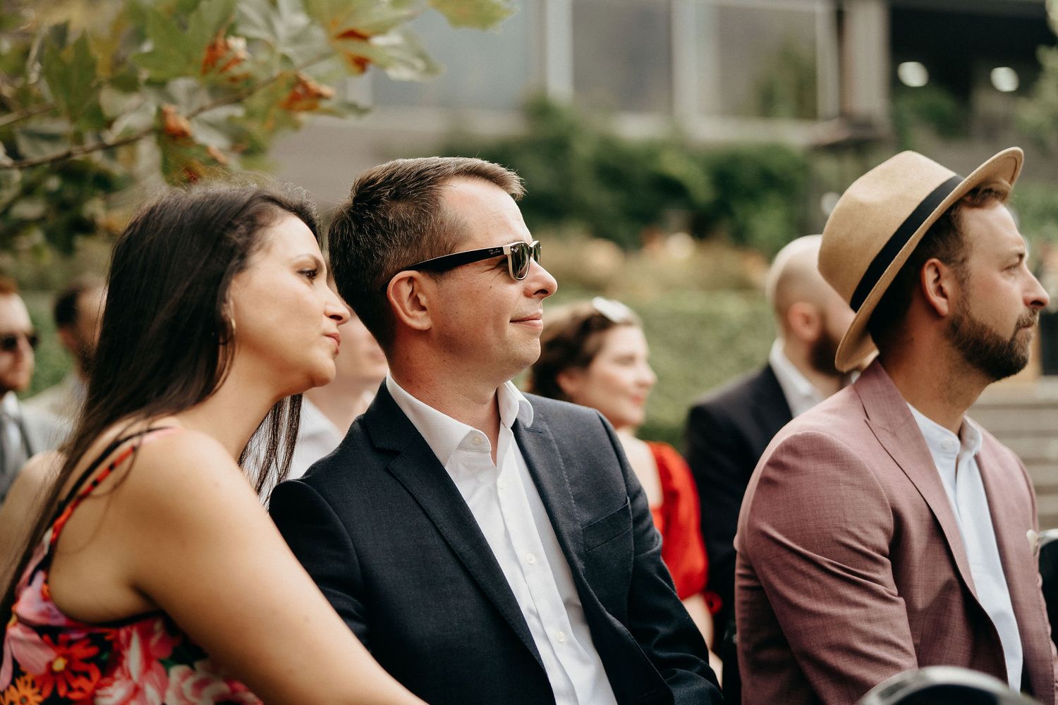 Wedding guests sit outdoors during a ceremony with foliage visible in the background.