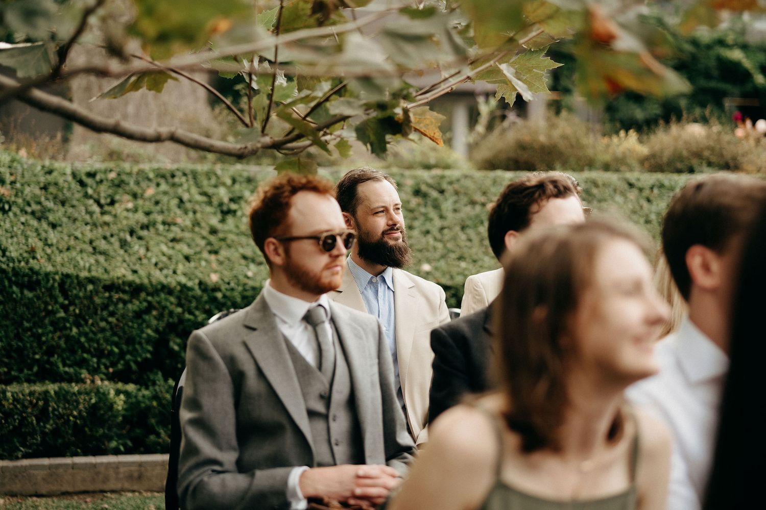 Wedding guests mingle outdoors in front of a lush green garden hedge during a sunny afternoon ceremony.