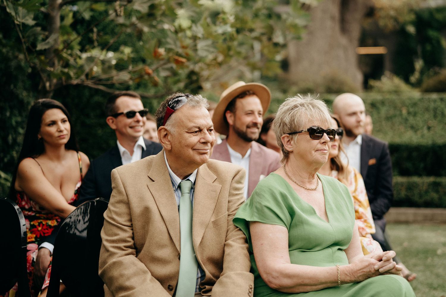 Wedding guests seated outdoors smile and enjoy the ceremony on a sunny summer day.